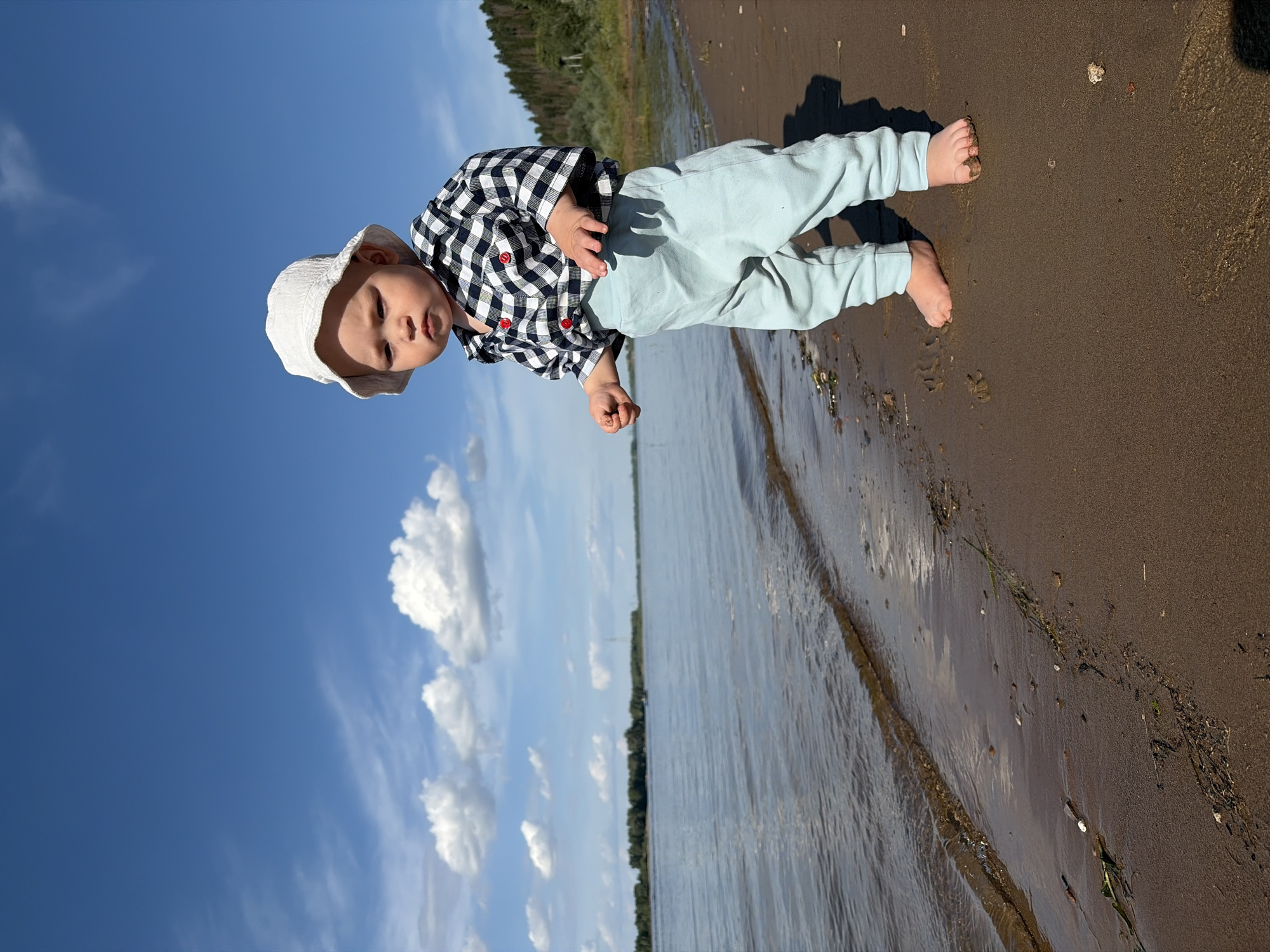 A young child, wearing a white bucket hat and a black and white checkered shirt with red buttons, stands on a sandy shore. The child is also wearing light blue pants. The child is facing slightly to the left of the camera, with their right hand outstretched. They appear to be looking down. The background shows a wide body of water, likely a river, with a line of trees and a hill on the opposite bank. The sky is blue with scattered white clouds. The scene appears to be outdoors in a natural setting, possibly near the city of Olkhovka, Russia, given the provided context. The sandy shore has some debris and footprints. The water has small waves. The lighting suggests it is daytime with bright sunlight.