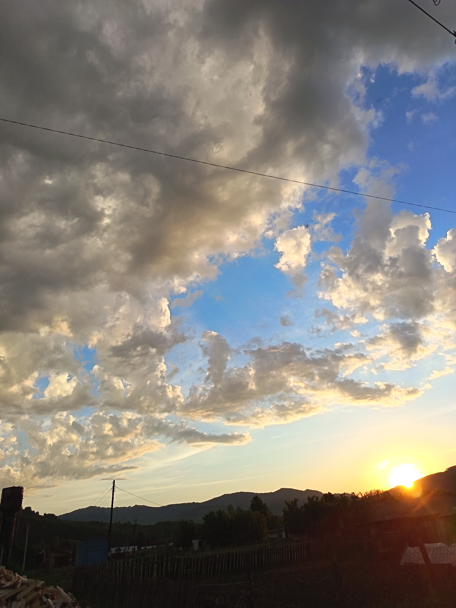 The image depicts a rural landscape at sunset in Soloneshnoye, Russia. The sky is filled with dramatic clouds, ranging from dark gray to bright white and tinged with golden hues from the setting sun. A power line stretches horizontally across the upper portion of the frame. The foreground shows a dark, silhouetted rural village with trees and a wooden fence. In the far right of the frame, the sun is setting behind a hill, casting a bright glare and orange light. To the left, a water tower and a pile of firewood are visible. The overall impression is one of a peaceful, yet dynamic, rural evening.