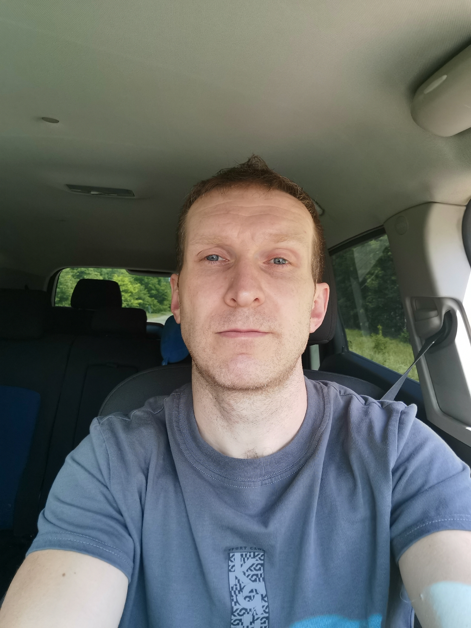 A man is sitting in the driver's seat of a car, looking directly at the camera. He has light skin, short brown hair, and blue eyes. He is wearing a grey t-shirt. The interior of the car is visible, including the headliner, seats, and seatbelt. Outside the car, trees and greenery can be seen through the windows. The scene appears to be in a rural or suburban area.
