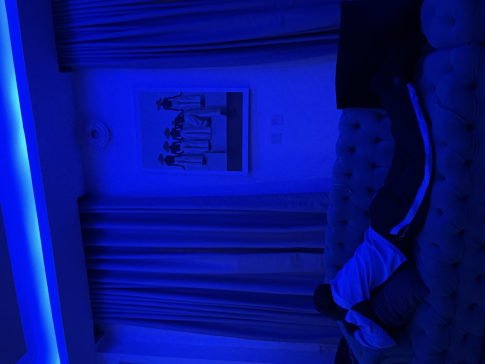 A person lies on a tufted blue sofa in a dimly lit room, illuminated by bright blue accent lighting from the ceiling. The person is wearing a white hoodie and dark pants with a white stripe down the side, and is lying on their side with their head resting on a pillow. To the right of the person, on the sofa, is a dark pillow. Behind the sofa, blue curtains hang on the wall. On the wall above the sofa, a framed artwork depicts five silhouetted figures wearing wide-brimmed hats and white outfits. The room appears to be a living room or entertainment space. The image is undated and unlocated, but the prompt specifies it is in Ado, Nigeria.