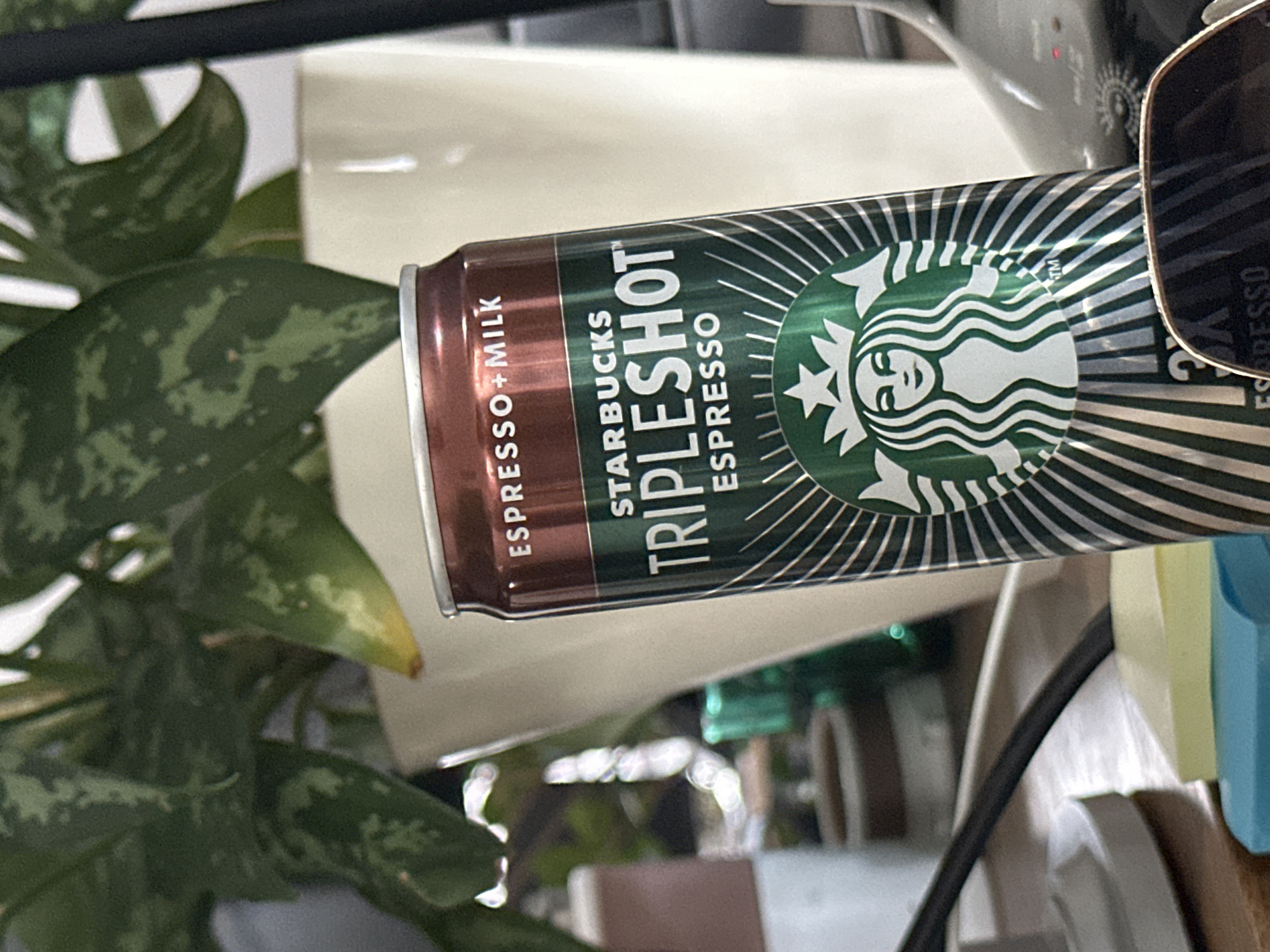 A Starbucks Triple Shot Espresso can is in the foreground, angled towards the viewer. The can is primarily green and silver with the Starbucks logo and text indicating "TRIPLESHOT ESPRESSO" and "ESPRESSO+MILK". Behind the can, a green leafy plant is visible, with some leaves yellowing. In the background, there are two white cylindrical objects, possibly vases, and a blurred object on the left. The lighting suggests an indoor setting. The image does not contain any information that would allow for the determination of the location being the city of Khimki, Russia.