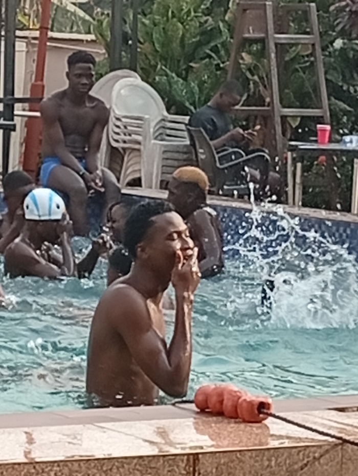 People gather around a pool, with several individuals swimming or standing in the water. One man is in the foreground, near a floating orange pool divider, raising his hand to his mouth. Behind them, other men sit on chairs near the pool’s edge. Some are shirtless, while others wear dark shirts. A ladder stands against lush green plants. The scene appears relaxed and recreational.