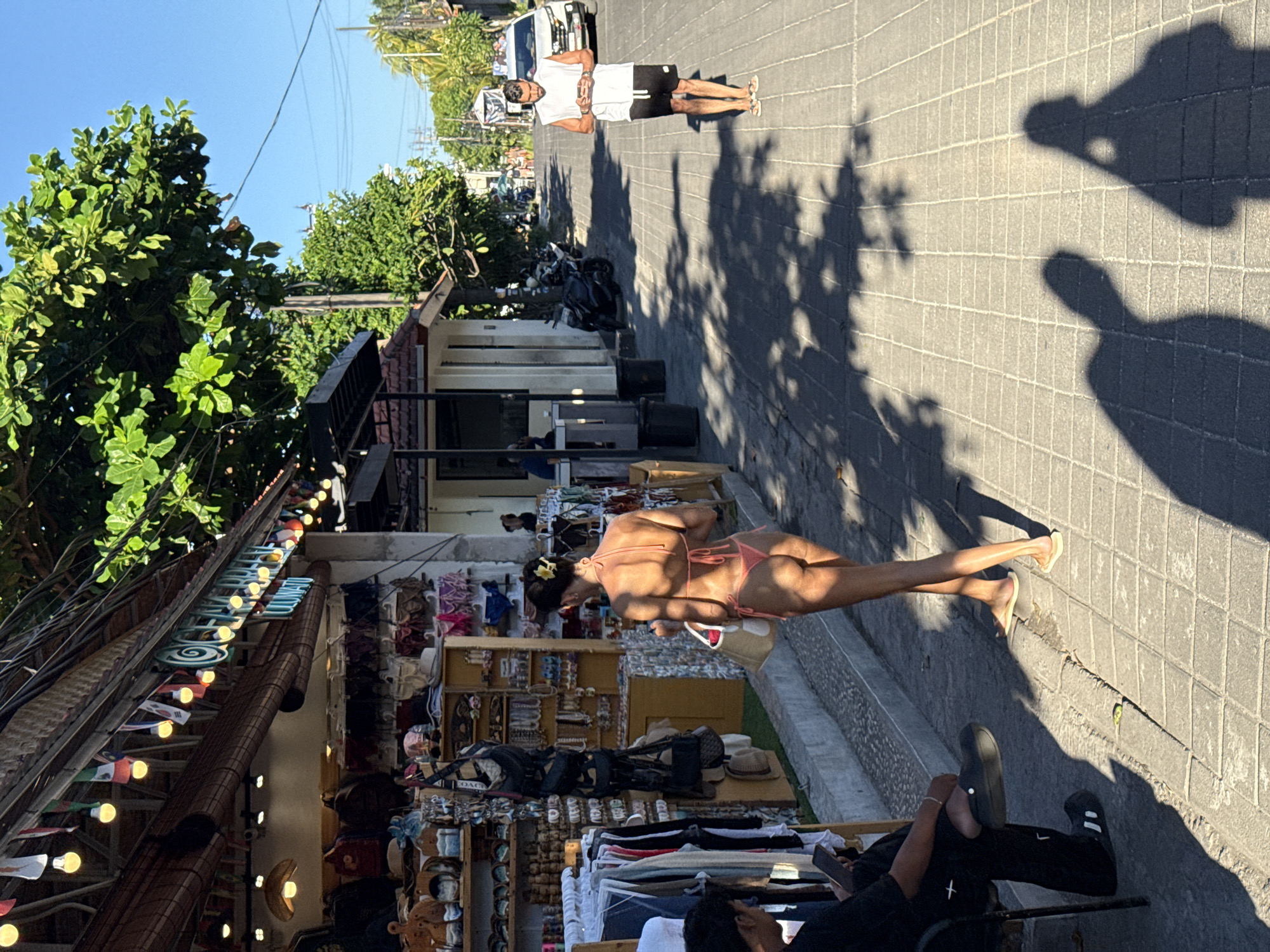 This vertically oriented image captures a bustling, sunlit street scene in Kuta Selatan, Indonesia, likely a commercial or market area. The time of day is indicated by strong, elongated shadows cast across the tiled pavement, suggesting either morning or late afternoon. The sky is clear and blue.
On the left side, a series of open-front shops and stalls line the street, displaying various goods such as clothing, bags, hats, and other accessories. Small, illuminated bulbs are strung along the shop awnings. In the immediate foreground, a person is seated, their feet in black sandals visible.
In the midground, a woman in a pink bikini top and bottom walks along the pavement, looking towards the shops. She has a flower in her hair and is holding what appears to be a white bag. Further down the street, a man in a white t-shirt, black shorts, and flip-flops stands near a white vehicle. He has his hands clasped in front of him. Motorcycles are also parked further along the street.
The pavement is made of square or rectangular tiles, and the road surface appears to be similarly paved. Overhead power lines are visible against the clear sky, and lush green tropical trees flank the street. Inside one of the shops, a bag with the brand name "COACH" is clearly visible. The scene depicts a casual, lively atmosphere with people engaged in daily activities like walking and shopping.