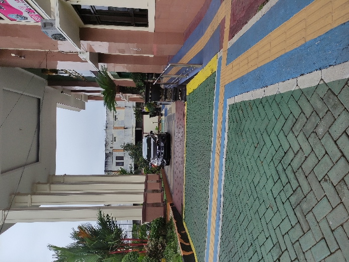 This outdoor image, taken in Kepanjen, Indonesia, is oriented vertically, showing a vibrant paved area alongside buildings under an overcast daytime sky.

In the foreground, the ground is covered with colorful interlocking pavers. A prominent section features a green herringbone pattern, bordered by bold stripes of blue, yellow, and white. Further back, segments of red or maroon paving are also visible.

To the left, a multi-story building with brown walls and white trim dominates the scene. An air conditioning unit is affixed to an upper section. On one of its upper white walls, a sign with pink lettering is visible, clearly displaying "SALON" and "HAIR MAKE UP," suggesting a beauty salon. Further to the left and slightly recessed, another building with large white columns and a decorative ceiling is partially seen, framed by green foliage and small palm-like plants.

A dark-colored car, appearing to be a compact SUV or sedan, is parked on the paved surface in the midground, facing away from the viewer. It is positioned on a section of the blue-striped paving. Beyond the car, various multi-story buildings with lighter facades are visible against the bright, featureless sky. Scattered green bushes and small trees are present throughout the scene, adding natural elements to the built environment.

There are no people visible, and no specific activity is taking place; the scene is static. The bright, uniform sky indicates it is daytime, likely an overcast day.