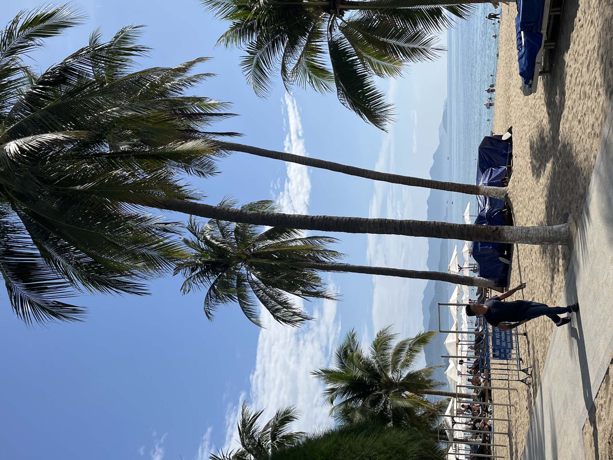 In Nha Trang, Vietnam, a man walks along a paved path bordering a sandy beach. The beach is lined with palm trees, beach chairs, and umbrellas, some covered in blue tarps. In the distance, the blue ocean meets a hazy horizon with mountains. Several people are visible on the beach and swimming in the water. The sky is a clear blue with scattered white clouds.