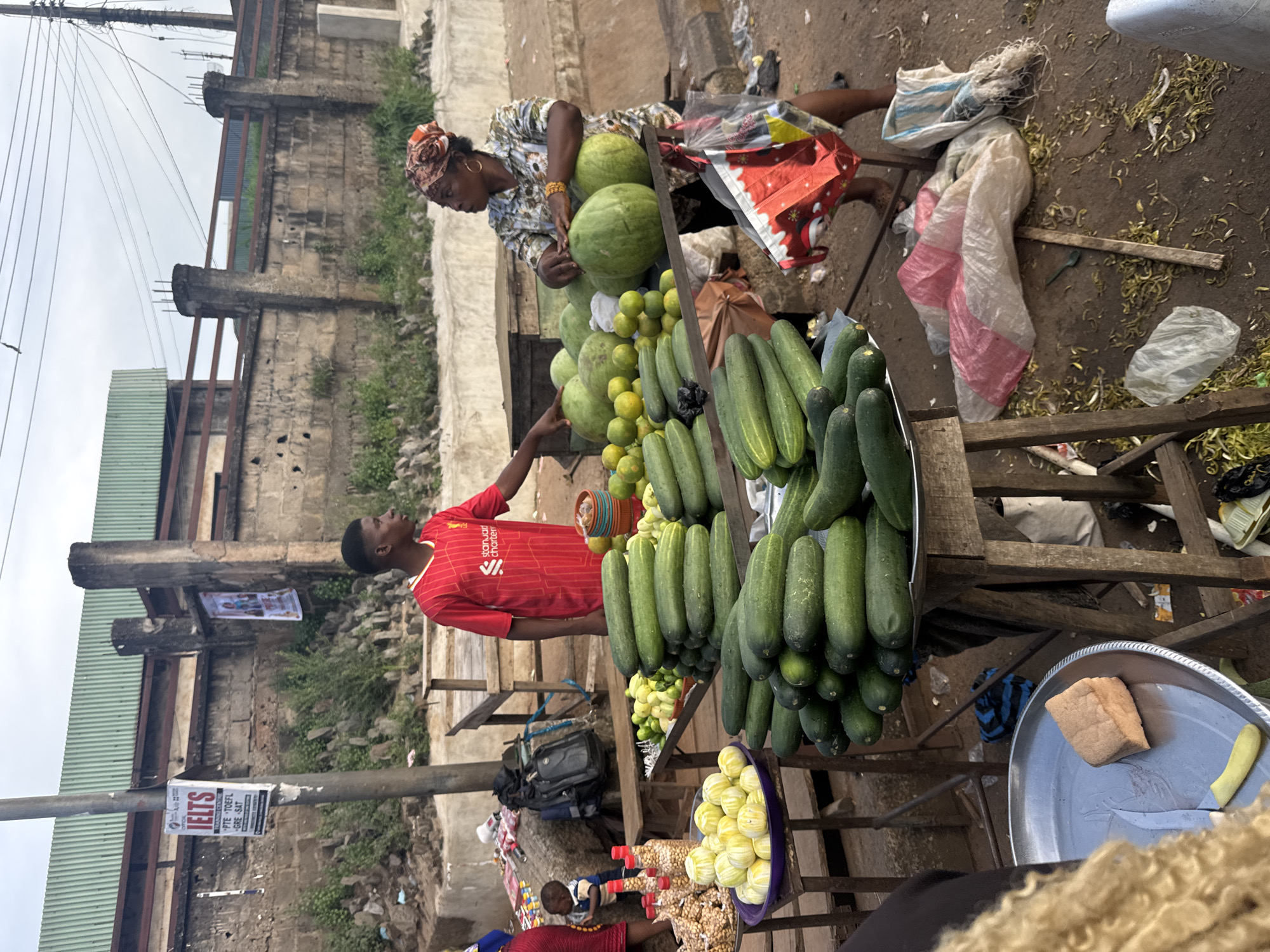 A man in a red shirt and a woman in a patterned top with a headscarf stand at a market stall. The man is pointing towards a pile of cucumbers, while the woman is arranging watermelons. Surrounding them are various produce, including limes, and other items. The scene appears to be an outdoor market with a backdrop of buildings and a cloudy sky, suggesting it might be daytime. The ground is dirt and debris. In the background, posters are visible on a wall, one of which advertises "IELTS PTE TOEFL GRE SAT". This setting is identified as Onitsha, Nigeria.