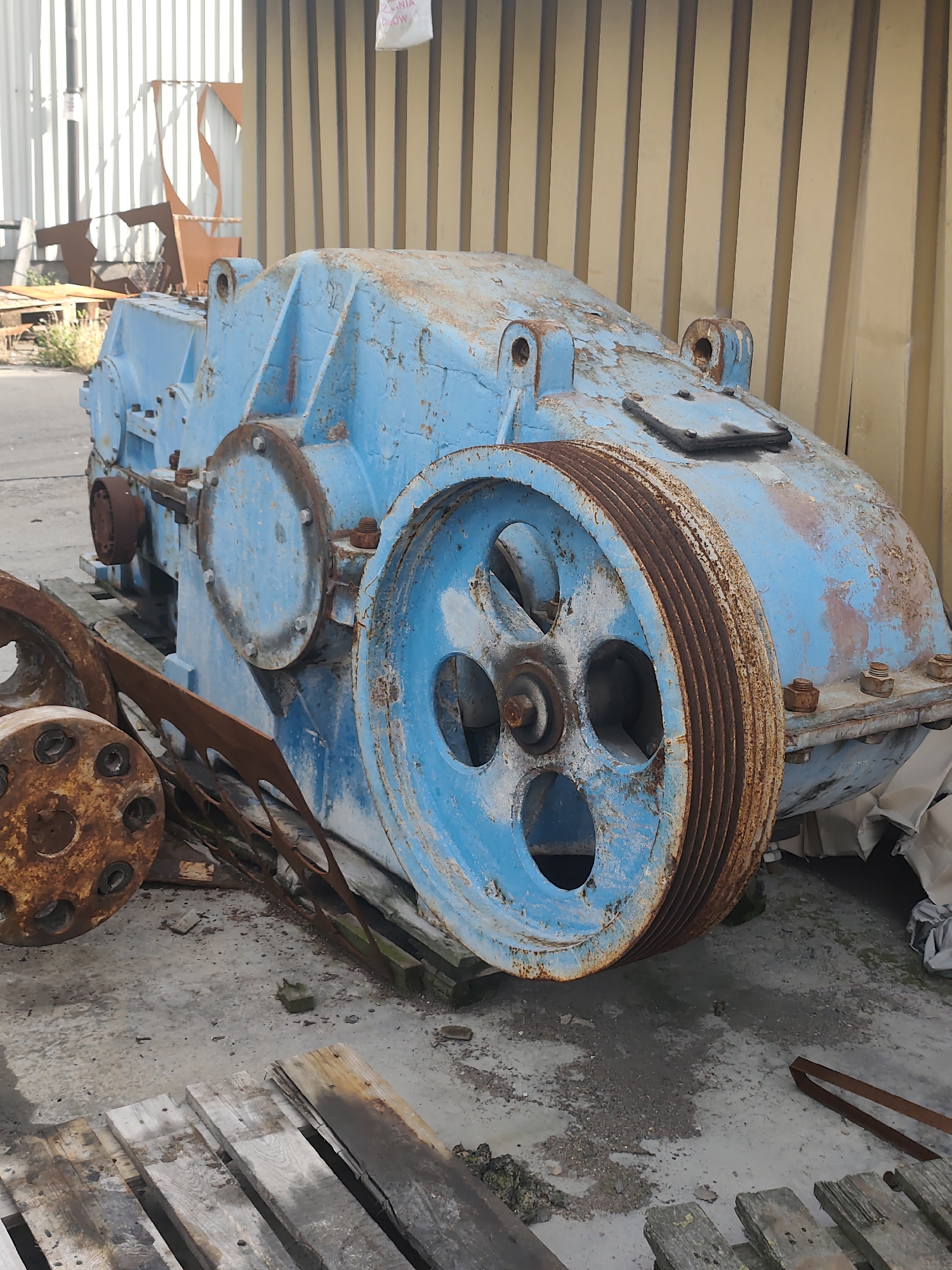 A weathered, industrial blue machine, possibly a large grinder or compressor, rests on a concrete surface in an outdoor industrial area. The machine exhibits significant rust and peeling paint, indicating exposure to the elements. It features a large, ribbed pulley wheel on its right side, also heavily rusted. To the left, another rusted wheel is visible. The background consists of a tall, corrugated metal structure with vertical panels, painted a dull yellow. Scattered around the machine are wooden pallets, some broken and worn, and other discarded metal objects. The overall environment suggests a scrapyard or an area for storing industrial equipment. The location is identified as Police, Poland.