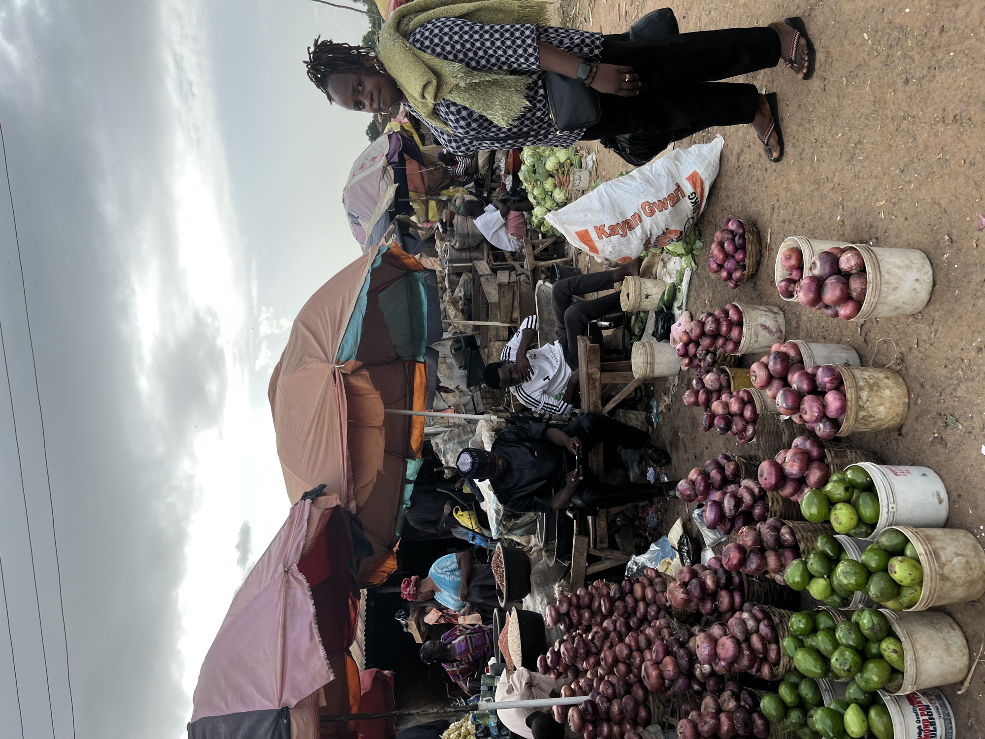 In Rafin Par, Nigeria, a market scene unfolds under a cloudy sky. The foreground features a woman standing, draped in a light green shawl over a black and white checkered shirt and black pants. She wears flip-flops. To her left, piles of red onions are displayed in baskets and buckets, alongside buckets of green avocados. Behind her, a large white sack with red lettering is visible. The middle ground shows several people, some seated, in a market stall area covered by colorful umbrellas. A man in a black traditional outfit is seated, looking at his phone, next to a pile of onions. Further back, more market stalls with various goods and people are visible. The ground is dirt.