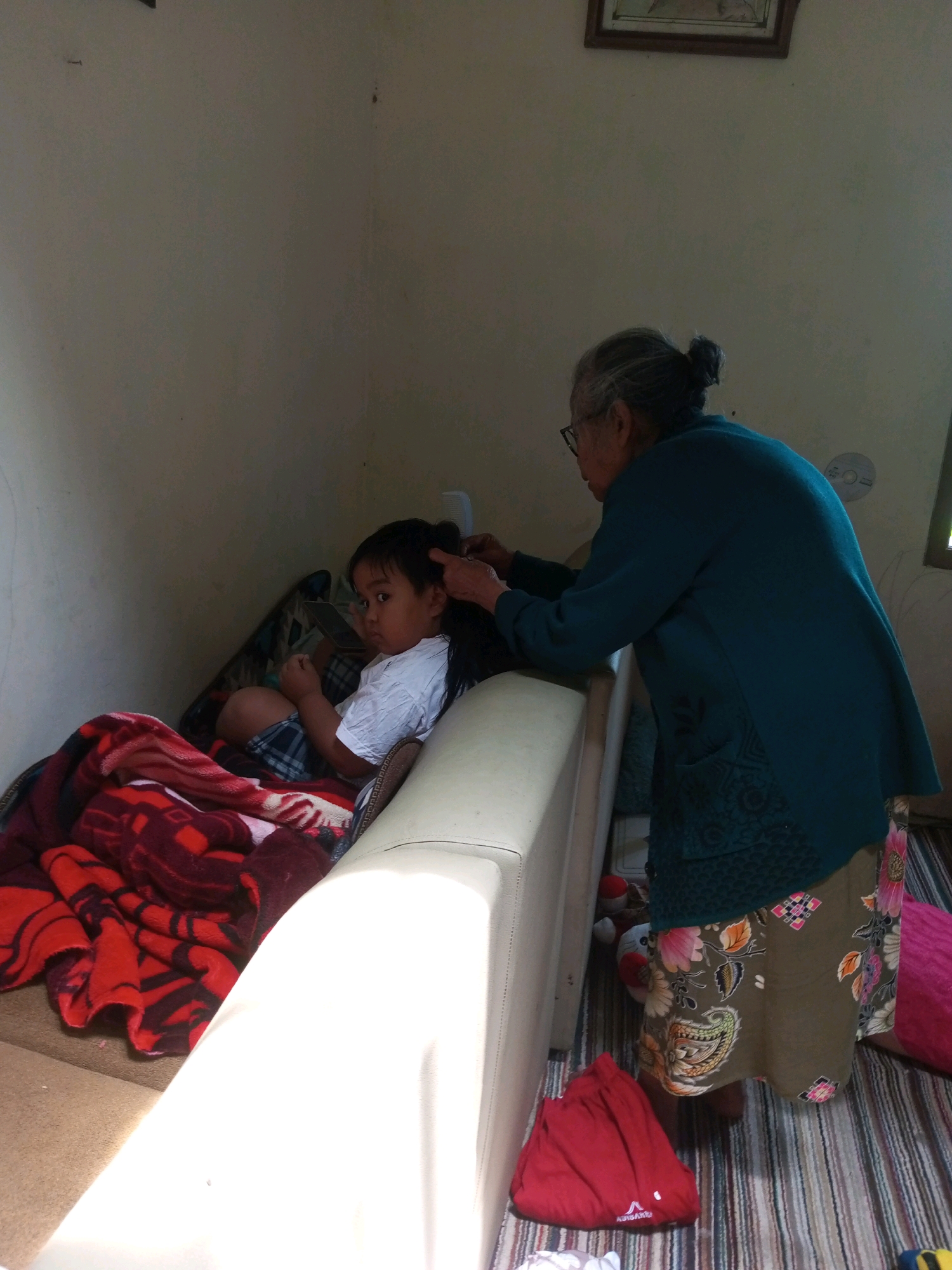 In a room, an elderly woman with her hair pulled back in a bun and wearing glasses combs the hair of a young child. The woman is wearing a teal cardigan over a patterned skirt. The child, wearing a white t-shirt and plaid shorts, is sitting on a sofa with blankets. The room appears to be lit by natural light, with a window visible in the background. The overall scene suggests a domestic setting, possibly a home in the city of Krasak, Indonesia.