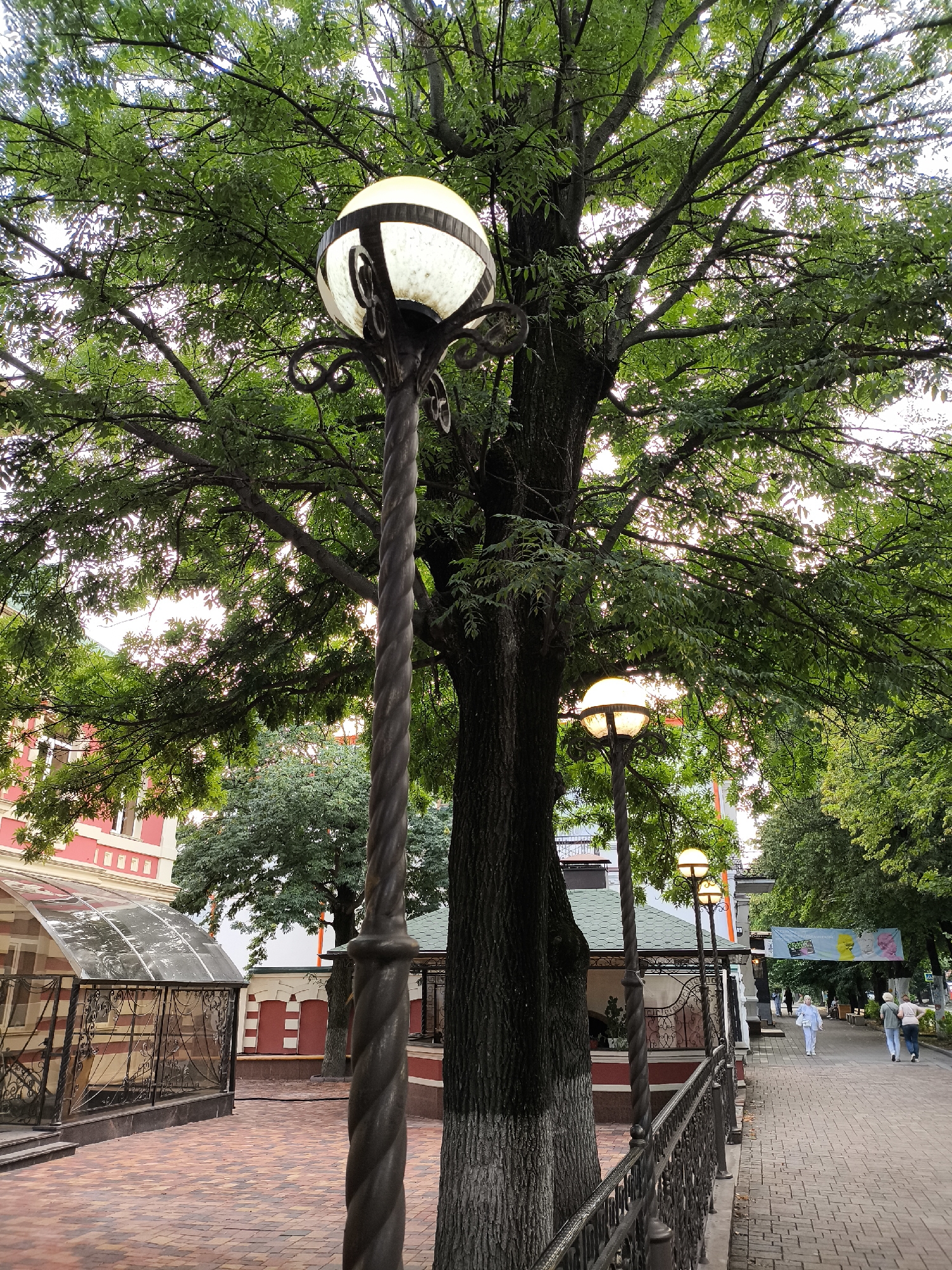 The image shows a tree-lined pedestrian walkway in Yessentuki, Russia. A large tree with lush green foliage dominates the foreground, its branches extending over the walkway. Several ornate streetlights with spherical glass globes are visible along the path. The walkway is paved with brick-like stones, and a decorative metal fence runs alongside it. On the left side of the walkway, there is a building with red and white accents, featuring a glass-enclosed structure. In the distance, several people are walking along the path. A banner hangs across the walkway in the background, featuring images of people. The overall scene depicts a well-maintained public space in Yessentuki.