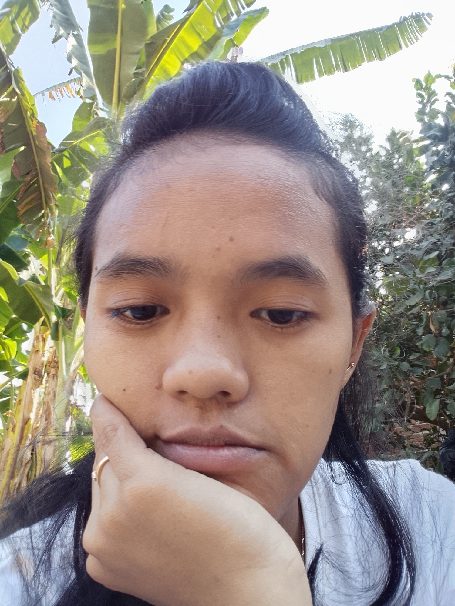The image is a close-up, selfie-style photograph of a young woman. She is outdoors, with a background of lush green banana leaves and trees. The lighting suggests it is daytime, possibly with direct sunlight. The woman's face is the primary focus; she has dark hair pulled up into a bun with some strands loose around her face. Her left hand is propped under her chin, and she is wearing a gold ring on her left ring finger. She appears to be wearing a white shirt. The environment, with the banana trees, suggests a tropical or subtropical setting. Based on the provided context, this scene is located in Kaponan, Indonesia.