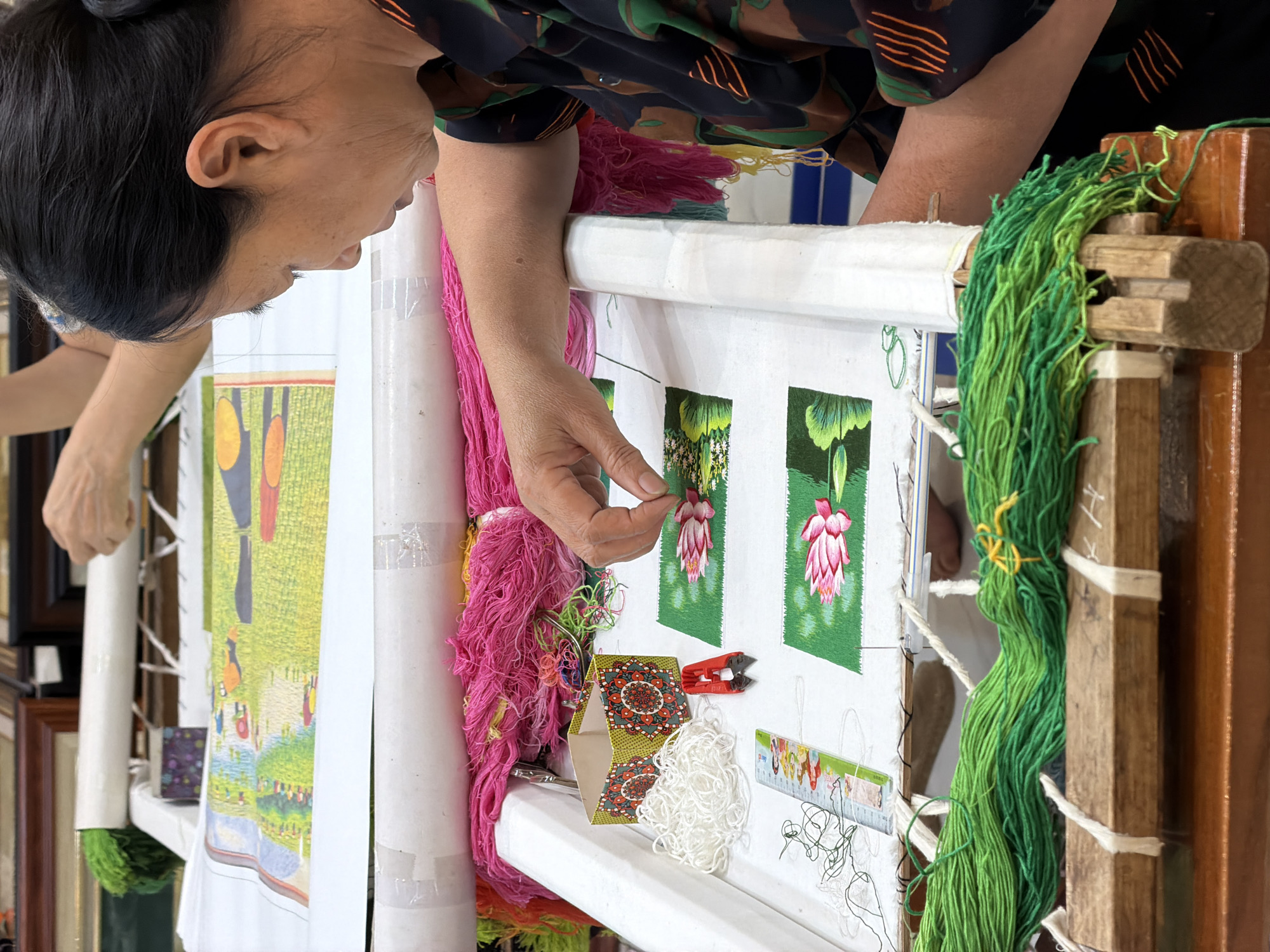The image captures a focused woman engaged in intricate hand embroidery in what appears to be a workshop or studio, likely in Hải Phòng, Vietnam. She is positioned over a large, upright embroidery frame, meticulously adding stitches to a piece of fabric.
The woman, with dark hair tied back, is seen in profile, her gaze directed intently at her work. Her right hand delicately manipulates a needle and thread, working on one of several small embroidered squares. Each square features a vivid pink and white fuchsia-like flower set against a deep green background. To her left, a larger, partially completed embroidery piece shows a more expansive scene with a light green field, abstract yellow and orange shapes, and smaller, indistinct figures. Another person's hand is visible in the background on the far left, suggesting collaborative work or another crafter nearby.
The embroidery frame is made of wood, with white fabric wrapped around its supports, holding the main canvas taut. Abundant bundles of vibrant threads are central to the scene. Large quantities of bright pink yarn hang from the left side of the frame, while various shades of green yarn are loosely wound around a wooden beam on the right. On a temporary ledge below the working area, small coils of white thread are visible, alongside a small red thread clipper and a decorative cardboard box with a mandala pattern. A ruler with colorful designs and faint markings also rests on the ledge.
The background is slightly blurred, revealing what might be framed pictures or mirrors, suggesting an artistic or craft-oriented environment. The overall impression is one of skilled craftsmanship and dedicated artistry, with a focus on traditional textile art. No prominent, readable text is visible on the embroidery itself or the surrounding objects beyond the faint markings on the ruler and wooden frame.