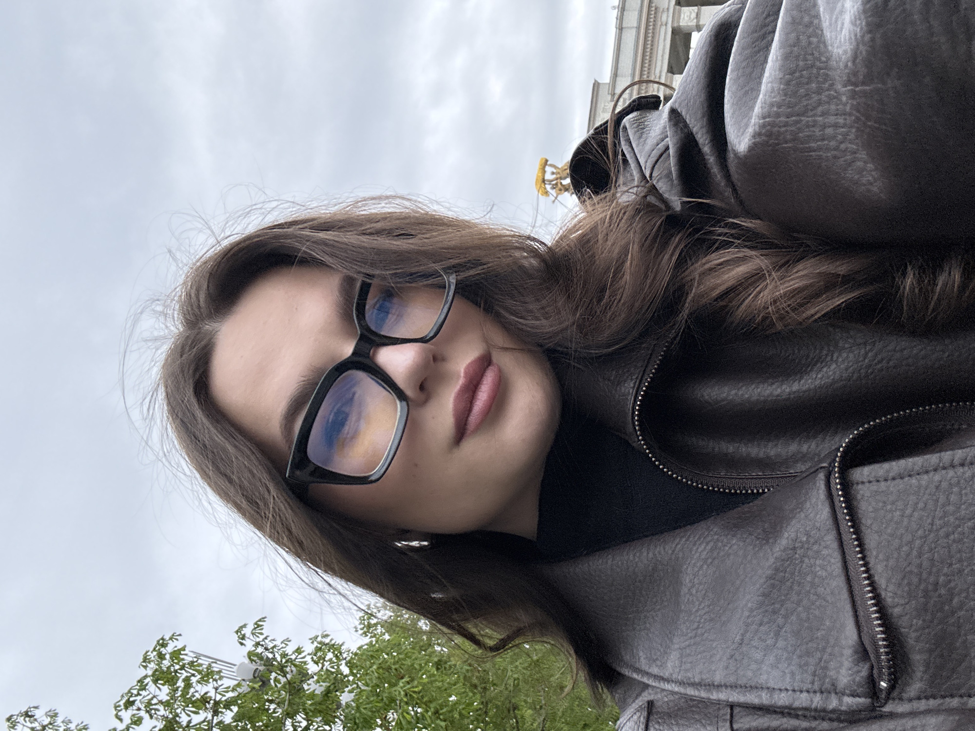 A young woman with dark hair and wearing black-framed glasses and a dark leather jacket poses for a selfie in Moscow, Russia. In the background is the cloudy sky above the city and the golden sculpture atop the Moscow State University building, along with the building itself. The tree is in front of the university. The woman has a neutral expression.