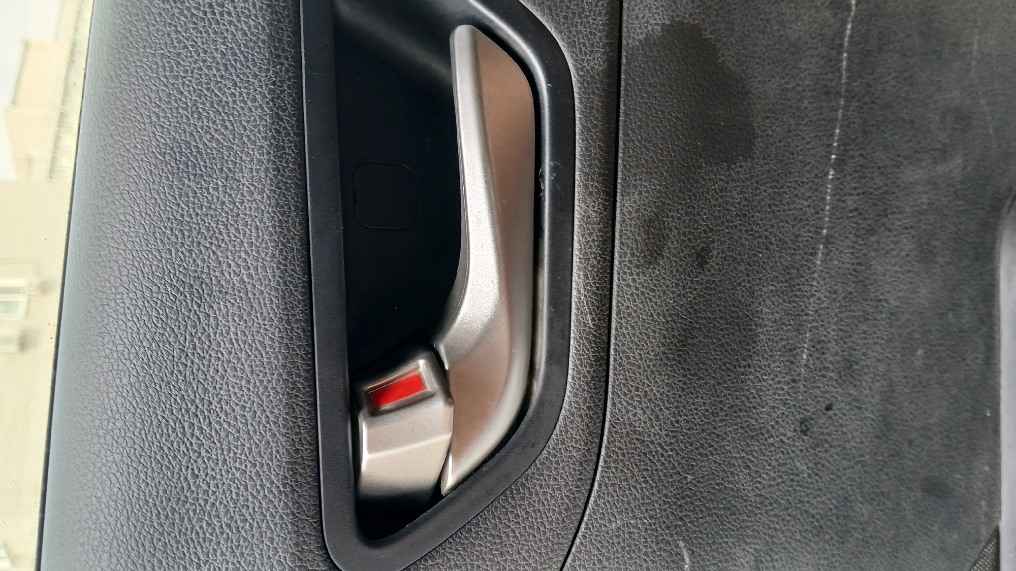 The image displays a close-up view of a car's interior door panel. A silver-colored door handle with a red accent is prominently featured. The door panel itself is made of black, textured material. The background is blurred, suggesting an outdoor setting with buildings visible. The image appears to be taken inside a car, likely a passenger vehicle, focusing on the door handle and surrounding panel. No people or specific actions are visible. The scene is static and details the state of the car's interior.