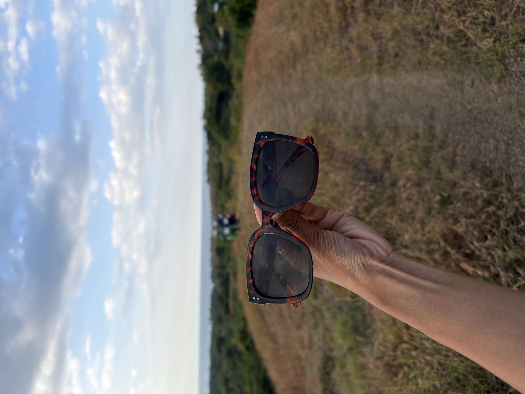 In the foreground, a person's left forearm and hand are visible, holding a pair of tortoiseshell-patterned sunglasses. The sunglasses are positioned slightly to the right of the frame, with their lenses facing forward. The background depicts a natural landscape, characterized by dry, grassy hills and a dirt path leading into the distance. In the far background, a body of water is visible, likely the ocean, meeting a hazy horizon under a sky filled with white and grey clouds. Further into the landscape, two individuals can be seen walking on the path in the distance. The scene suggests an outdoor setting, possibly a hiking trail or a vantage point overlooking the coast. The overall impression is one of a serene, natural environment.