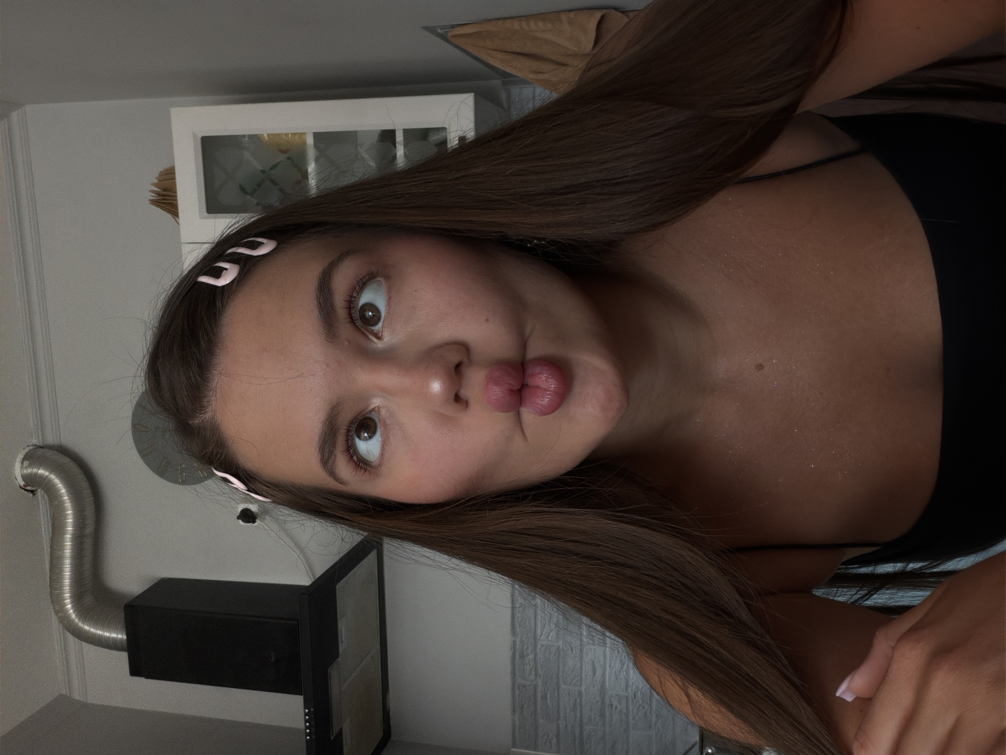 A close-up, low-angle, indoor, selfie-style shot depicts a young woman with long, dark brown hair, pulled back on the sides with two light pink hair clips. She is looking upwards with her eyes wide, and her lips are pursed in a fish-like pout. Her makeup includes winged eyeliner and blush. She is wearing a black camisole, and her collarbones and upper chest are visible, with a subtle shimmer of glitter.
In the background, a black kitchen range hood with a metallic flexible duct is mounted on a grey wall. To the right, a white cabinet with a glass door is visible. A beige towel hangs on the right side of the frame. The overall environment suggests a domestic kitchen setting. The image was taken in Sirius, Russia.