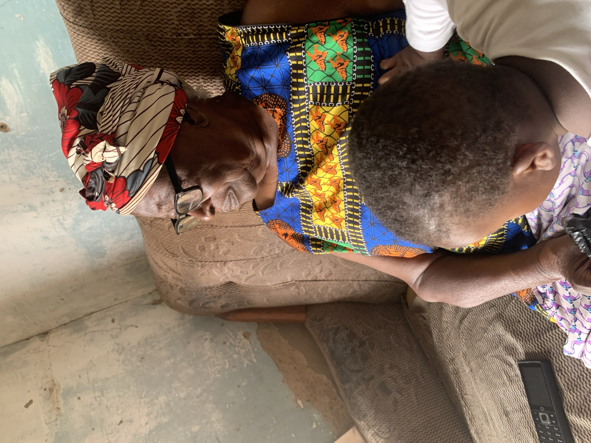 In Sabon Tasha, Nigeria, an elderly Black woman wearing glasses and a colorful headwrap sits on a brown armchair. She is dressed in a vibrant, patterned sleeveless dress. A toddler, with dark curly hair and wearing a white shirt, is positioned in front of her, facing away from the camera. The toddler's hand is resting on the woman's arm. A black remote control is visible on the striped fabric of the armchair next to the woman. The background reveals a wall with peeling paint in light blue and beige tones.