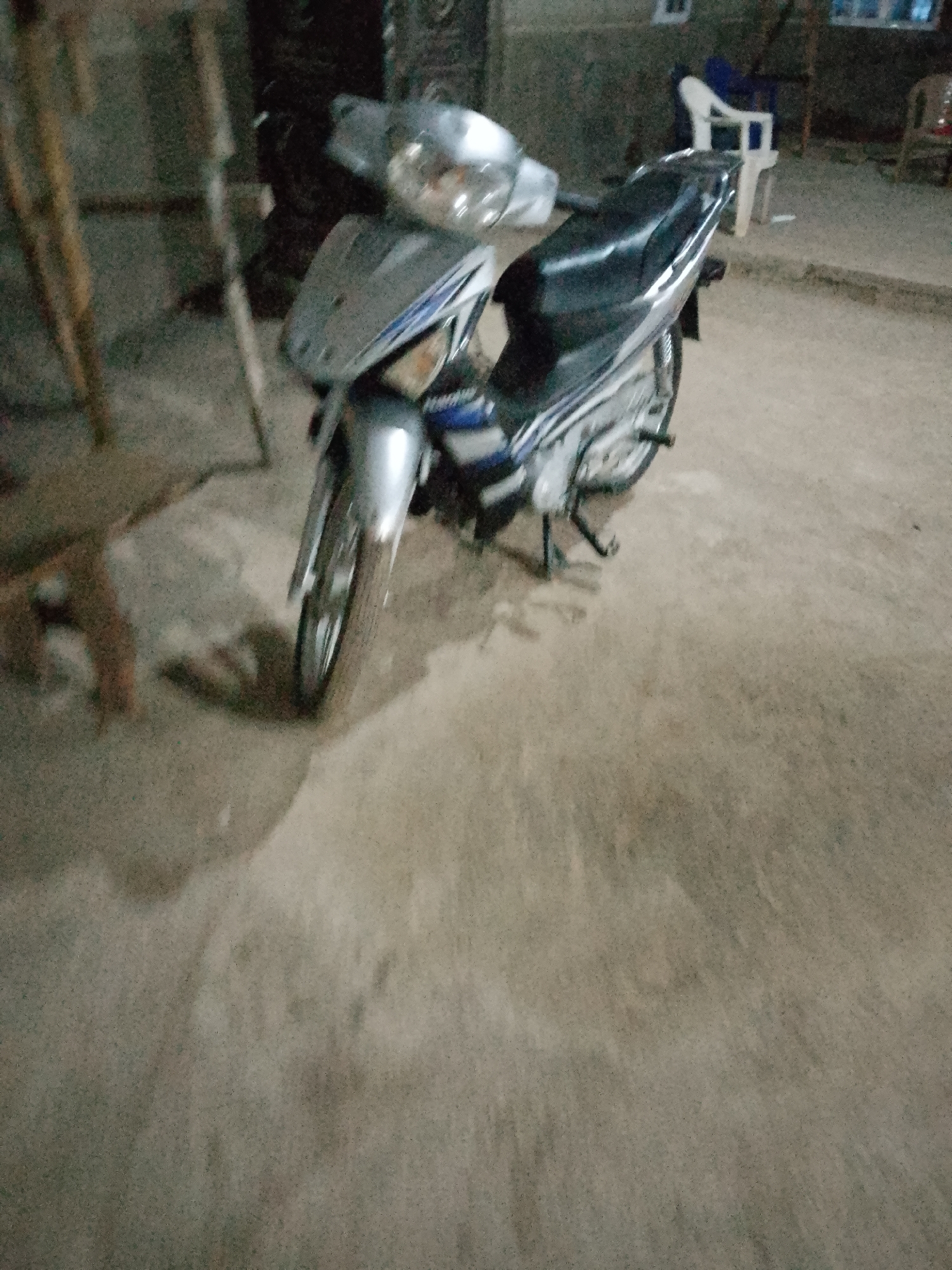 An overhead medium shot shows a silver and black motorcycle parked on a dirt ground. The motorcycle is facing left, with its front tire closest to the viewer. The motorcycle has a silver front fairing with blue and white accents. The seat is black. The background is blurred and appears to be an outdoor area, possibly a street or yard, with some wooden structures and furniture visible. The overall lighting is dim, suggesting it might be evening or nighttime. The location is identified as Serti, Nigeria.