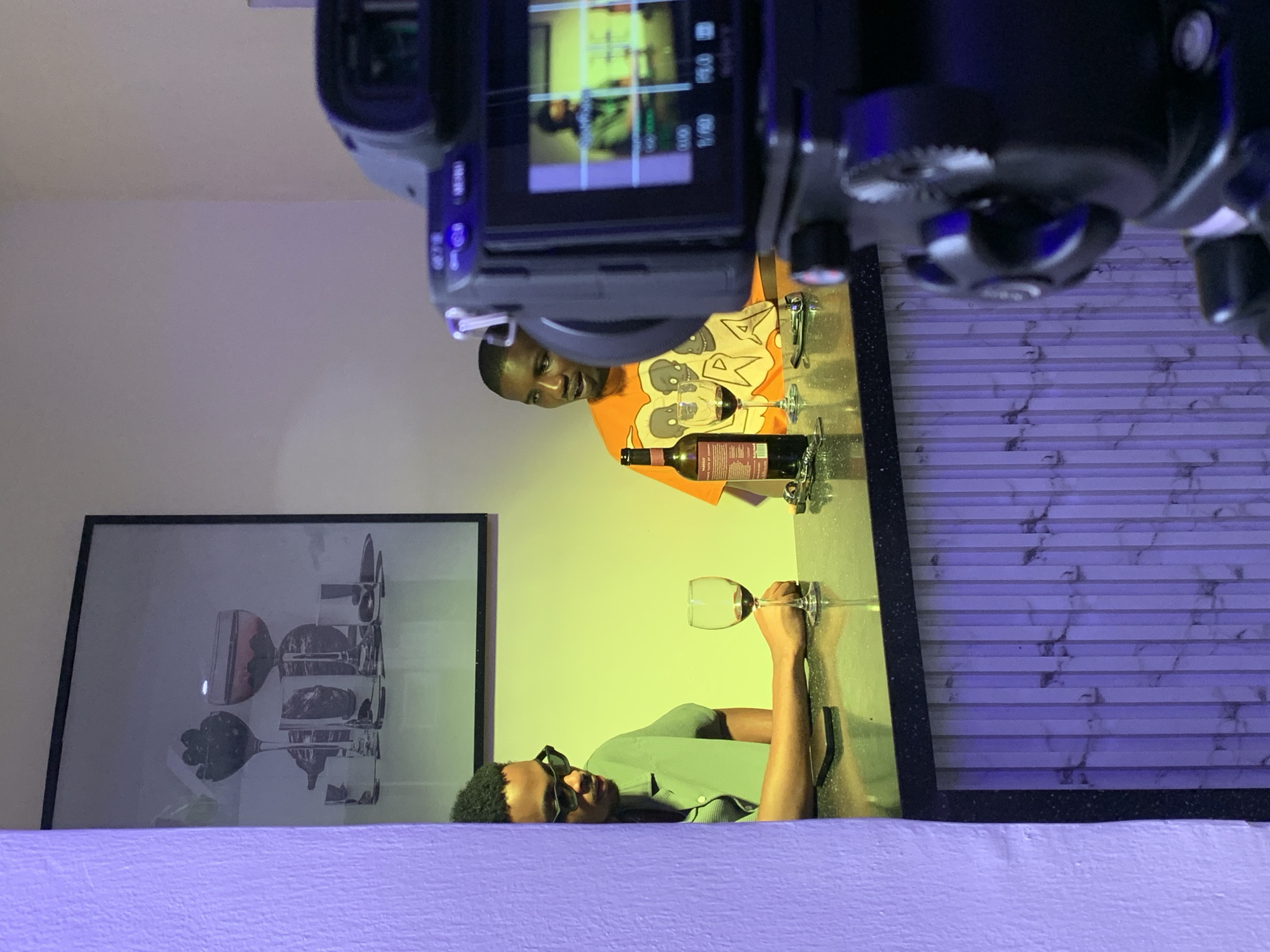 An indoor scene in Moba, Nigeria, depicts two adult males seated at a dark, reflective countertop, being filmed by a foreground camera. The man on the left, wearing black rectangular sunglasses and a light green collared shirt, sits with his left arm resting on the counter. A black smartphone and a wine glass containing red liquid are positioned in front of him. He is oriented towards the right. The man on the right, wearing an orange t-shirt with a graphic design, has a dark wine bottle with a red and white label, and another wine glass containing red liquid, in front of him. He is oriented towards the left, appearing engaged in conversation. The background wall is illuminated with a yellow-green light, featuring a large framed picture depicting stylized glassware. The immediate foreground wall to the left and the base of the counter exhibit vertical ribbed panels with a marble pattern, illuminated by a purple light. A black video camera, positioned in the lower right foreground, points towards the subjects, with its flip-out screen displaying a live feed of the man in the green shirt.