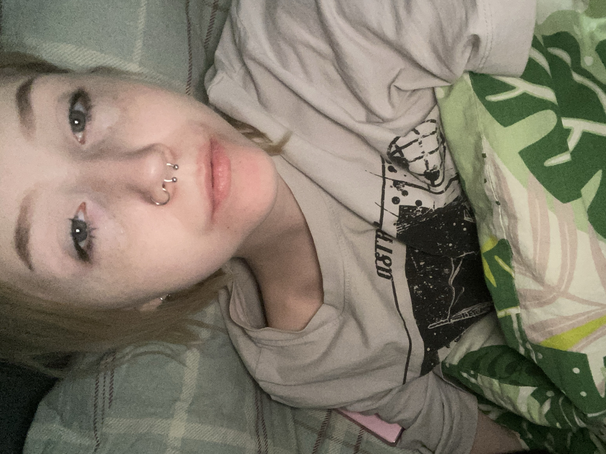 The scene depicts an individual in an indoor, domestic setting, consistent with a private residence in Izhevsk, Russia. A single person, appearing to be a young adult, is positioned lying down, looking directly towards the camera.
**People:**
One individual is present, with light-toned skin, short blonde or light brown hair, and dark eyes. Moisture is visible on the skin below both eyes, with slight redness around the lower eyelids. Freckles are discernible on the face.
**Subjects/Objects:**
The individual has multiple facial piercings: a silver horseshoe barbell and a silver ring in the septum, and a silver ring in the right nostril (viewer's left). A small silver hoop earring is present in the left earlobe (viewer's right). The person is wearing a light grey, short-sleeved t-shirt with a dark, abstract graphic print on the chest, which includes partial text (e.g., "AST R"). A pink object, consistent with a smartphone, is partially visible tucked under the person's left arm, resting on the bedding.
**Environment:**
The individual is partially covered by two layers of bedding. The uppermost layer, closer to the camera, is a duvet or blanket featuring a pattern of green and light-colored (possibly white or light pink/beige) tropical leaves. Beneath and behind the individual's head and left shoulder is a greenish-grey blanket or throw with a subtle, lighter plaid pattern. The lighting is dim, suggesting an interior space without bright direct light.
