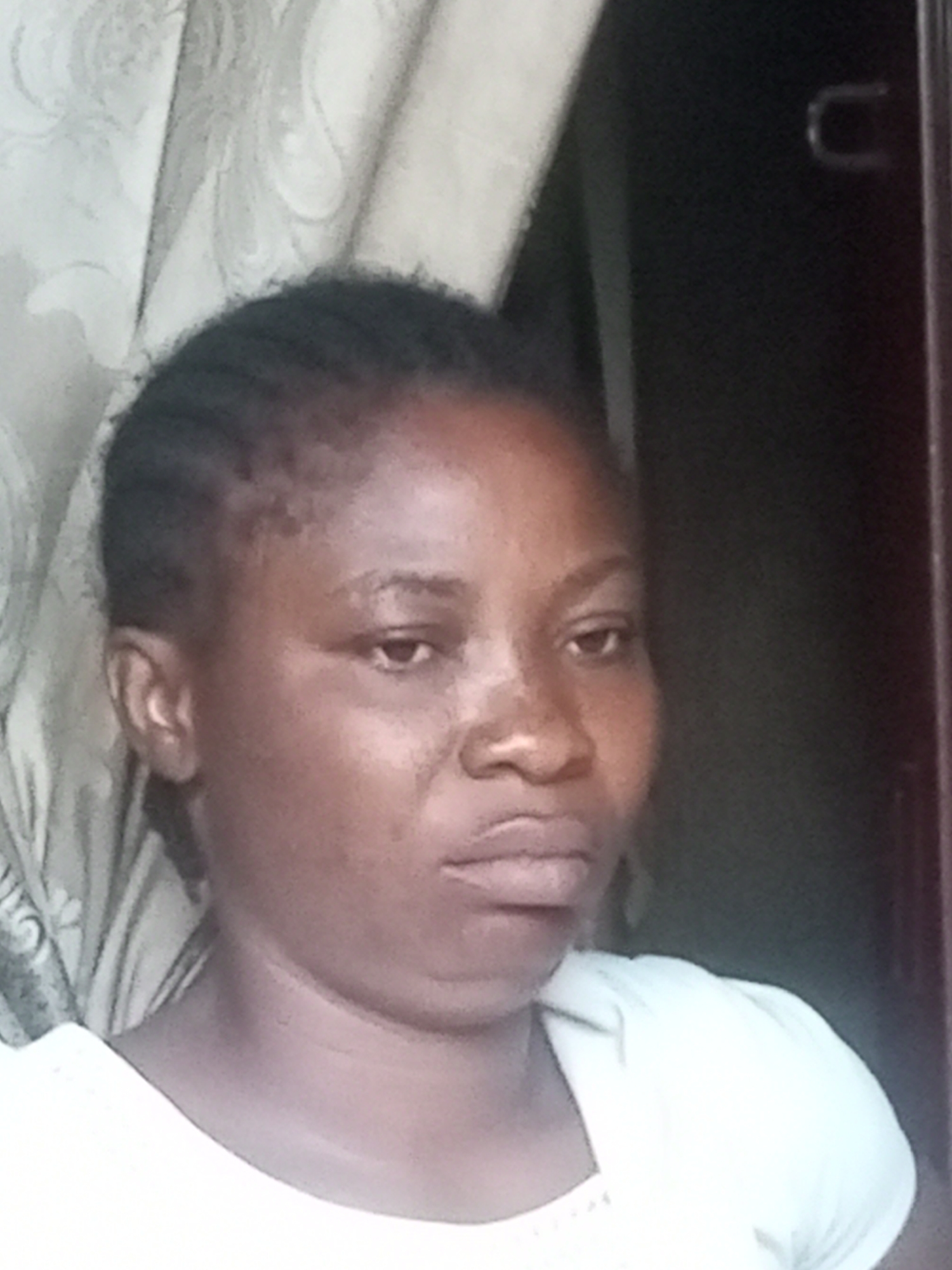 A medium shot, head-and-shoulders portrait of a young Black woman is captured from a slightly low angle. She is looking directly at the camera with a neutral expression. Her dark hair is styled in cornrows. She is wearing a plain white t-shirt. The background is softly blurred, with a patterned curtain visible on the left and a dark surface on the right, possibly a doorway or furniture. The lighting is diffused, casting gentle shadows on her face. There is no indication of the location being in Tempure, Nigeria, or any specific city.