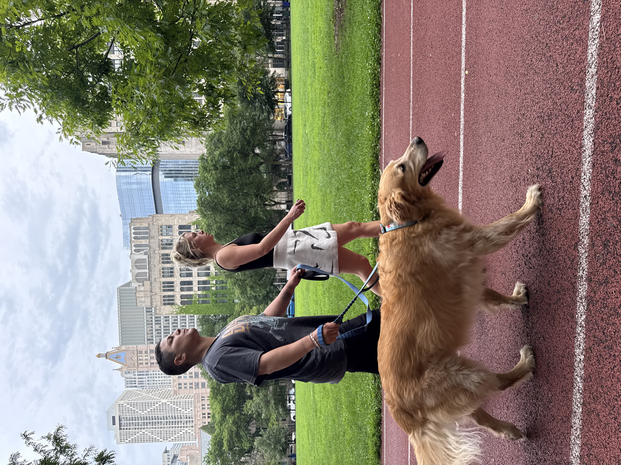 A golden retriever dog is being walked by two people on a red track with white lines, likely a running track in a park. In the background, a green lawn, trees, and several city buildings are visible under a cloudy sky. The man, on the left, is wearing a dark t-shirt and shorts, holding a blue leash with a braided handle. The woman, on the right, is wearing a black tank top and a white skirt, also holding the leash. The dog is panting, with its tongue out. The buildings in the background suggest an urban environment, possibly within the city of Chicago, United States of America.