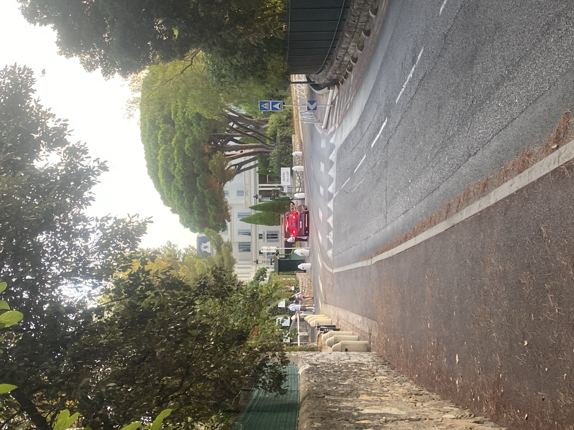 This image depicts a street scene in Antibes, France, near the Hotel du Cap-Eden-Roc. A red car is stopped at a pedestrian crossing on a paved road that curves to the right. To the left of the road is a stone wall topped with a green fence and a sidewalk lined with bollards. Beyond the sidewalk, there are trees and foliage. Across the street, a large white building, identified by a sign as "HOTEL DU CAP," is visible, framed by tall green trees. Two pedestrians stand on the sidewalk near the hotel entrance, and another individual stands by the hotel's gate. Traffic signs, including a pedestrian crossing sign and a directional arrow, are present near the road. The environment suggests a warm, possibly late afternoon, with sunlight filtering through the trees.