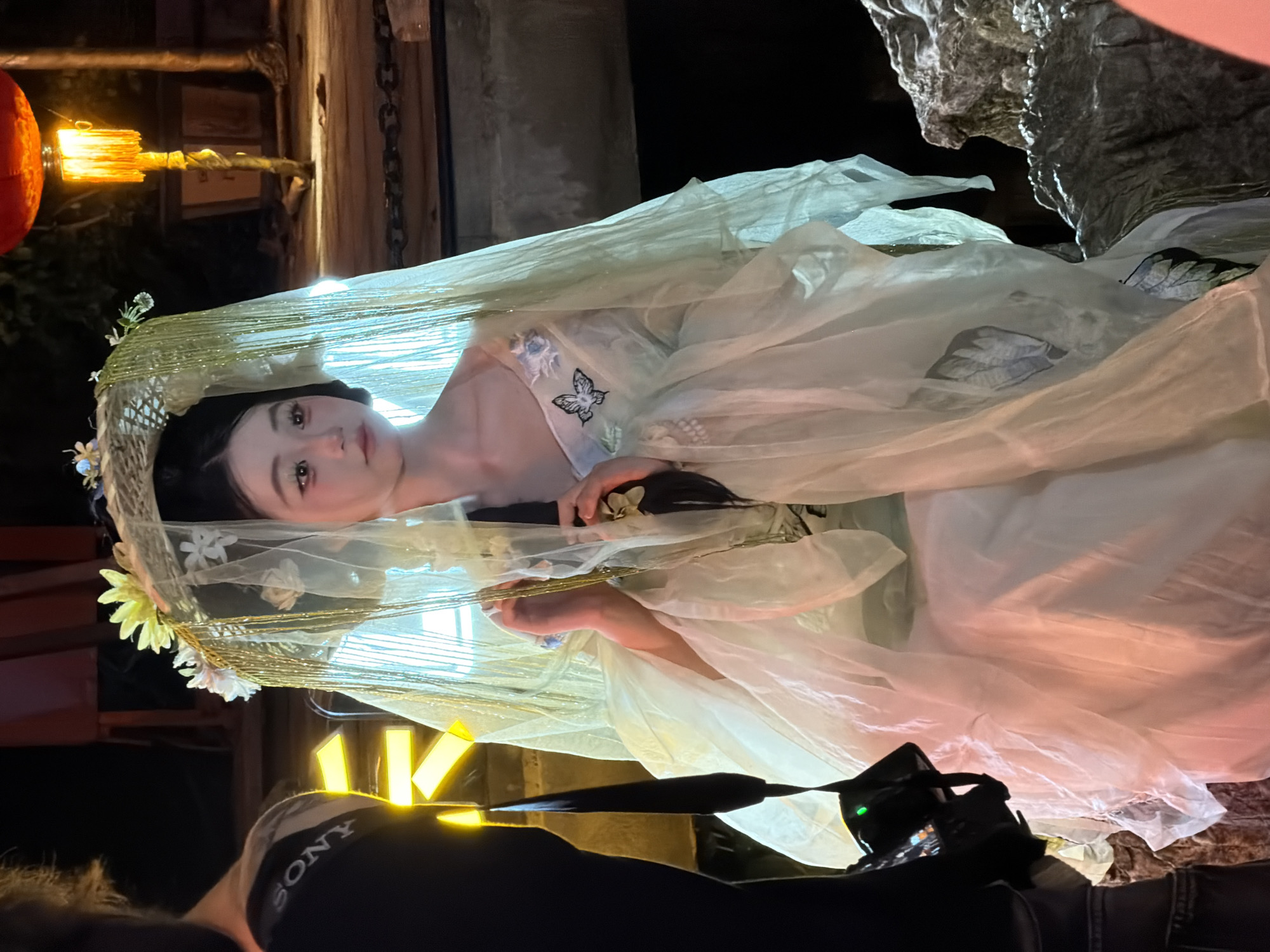 An individual, dressed in a pale green, ethereal gown with delicate floral and butterfly embellishments, is seated amidst a rocky outcrop at night. They wear an elaborate headdress adorned with flowers and shimmering gold tassels, partially obscuring their face. The individual's hair is styled in a braid, and their makeup is noticeable. To the left of the frame, a person wearing a black shirt with "SONY" printed on it stands partially visible, holding a camera. In the background, a red lantern emits a warm glow, illuminating a wooden walkway and potted plants. The overall scene suggests a staged photoshoot or artistic performance, possibly in the Yuzhong District of China, given the context.
