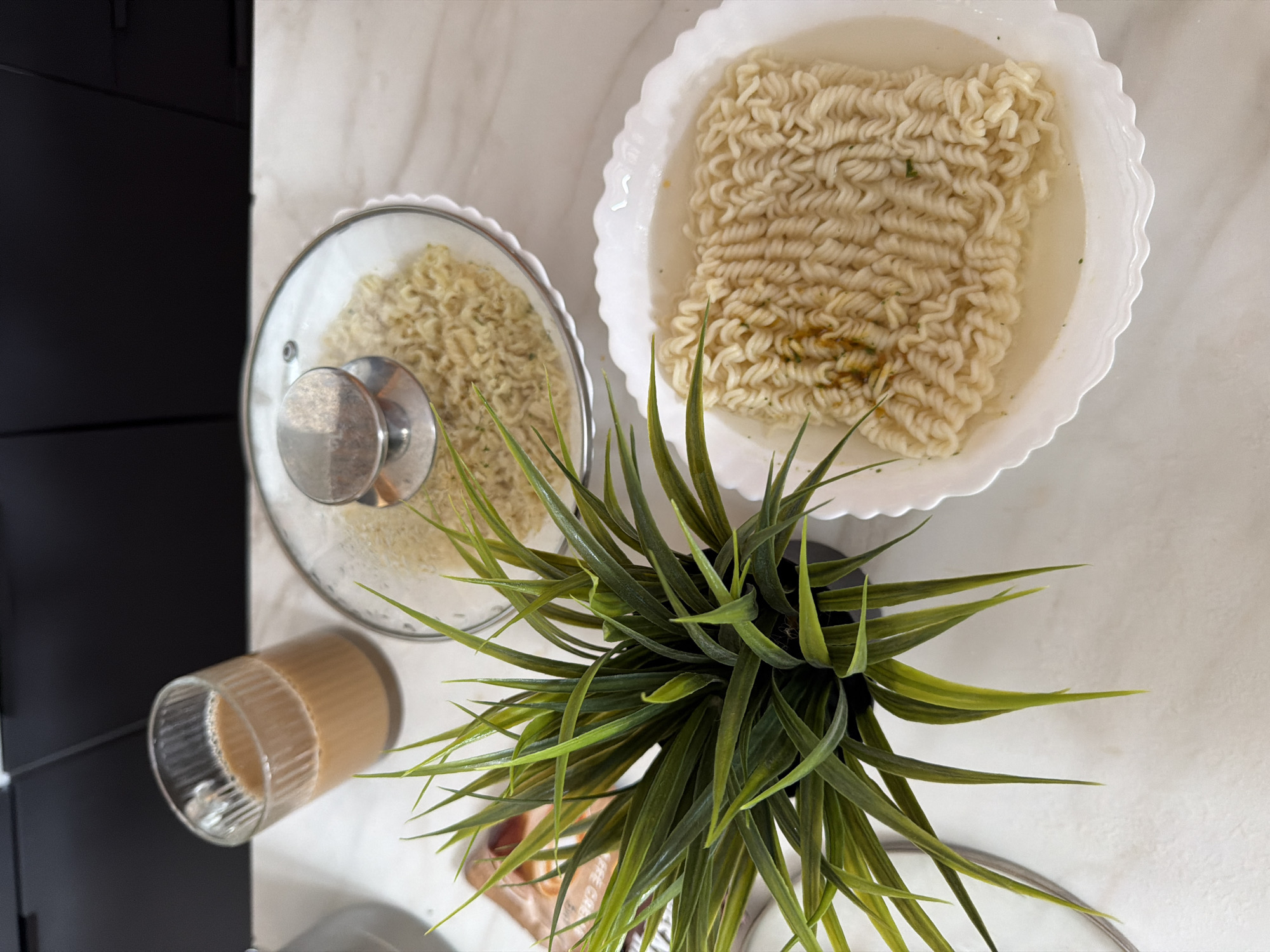 The image shows a close-up view of two bowls of instant noodles and a glass of tea on a white marble countertop. One bowl of noodles is covered with a clear glass lid with a silver handle. The other bowl of noodles is uncovered and partially submerged in broth. A small artificial green plant is positioned to the left of the bowls. In the background, there are black kitchen cabinets and a sliver of a silver appliance. The lighting is bright and appears to be from an overhead source. The scene is set in what appears to be a kitchen environment. There are no people or animals visible in the image. The location is described as being in the city of Семилуки, Russia, though this context cannot be visually verified from the image itself.