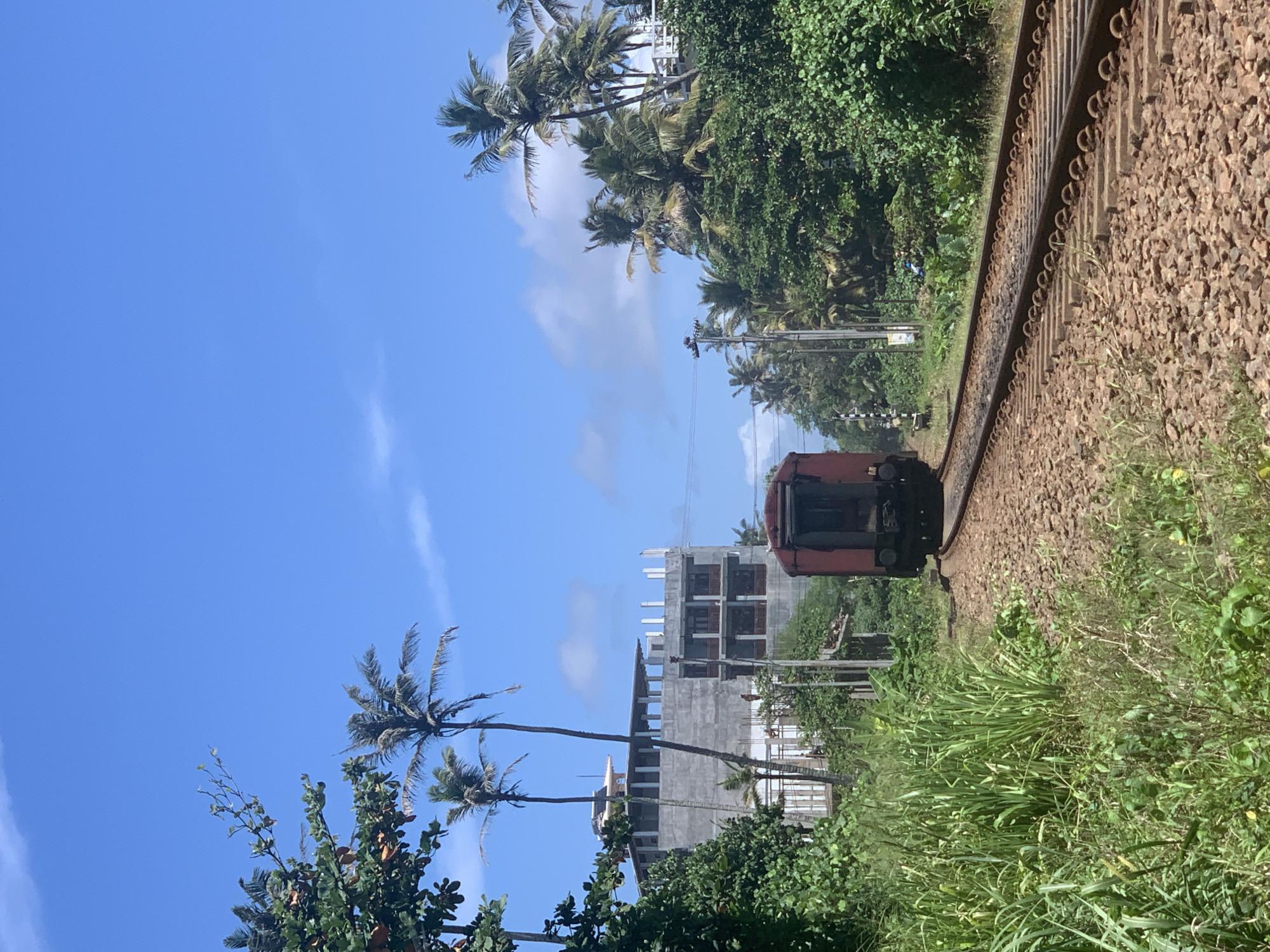 In Ahangama, Sri Lanka, a red train carriage is seen moving away from the viewer on a gravel railway track. The train is surrounded by lush green vegetation and palm trees. In the background, a multi-story building under construction with concrete walls and balconies is visible. The sky is a clear blue with a few scattered white clouds.