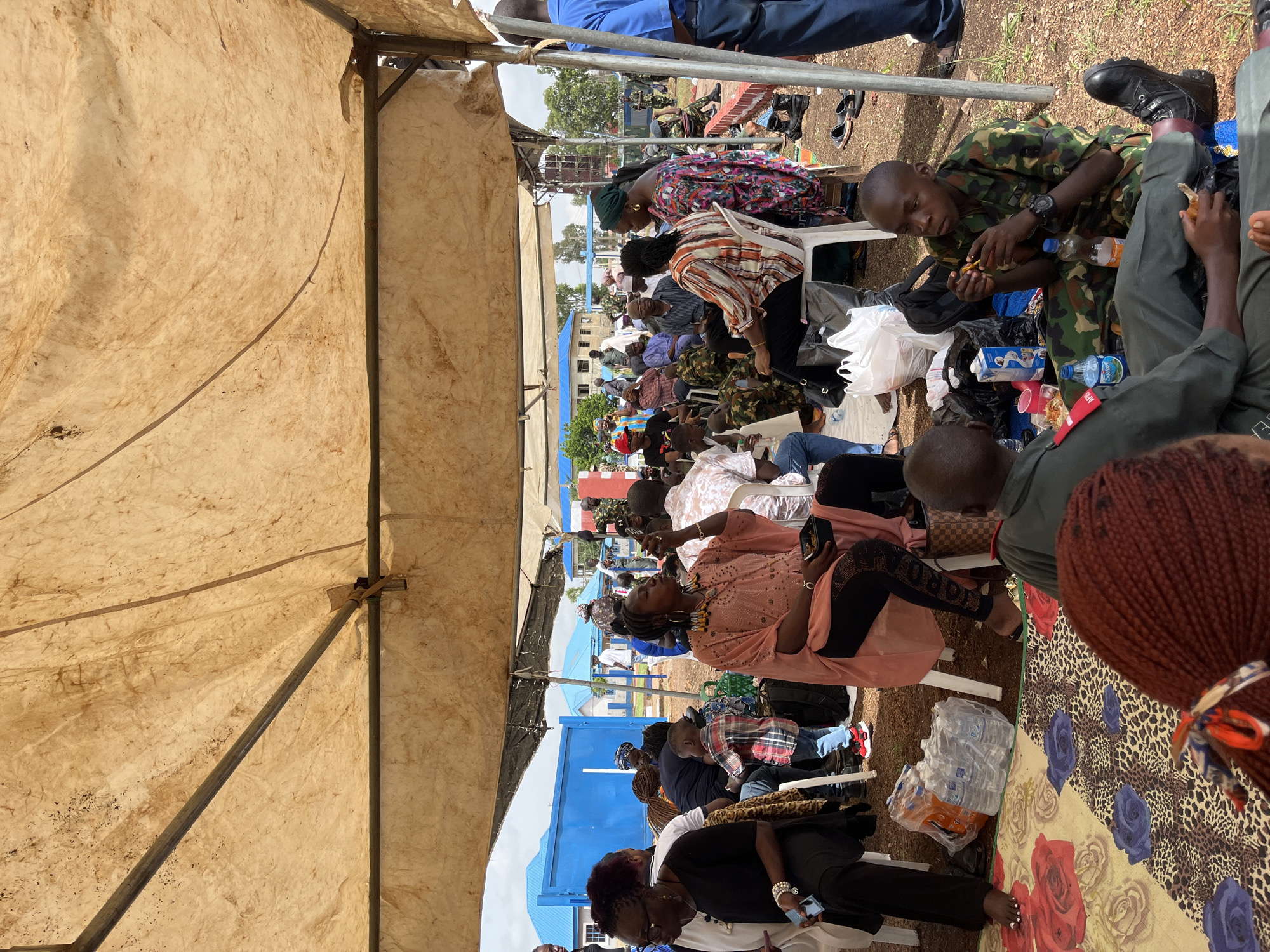 In Bar Jirgi Summa, Nigeria, under a large, weathered tan canvas tent, a gathering of people is observed. The tent is supported by metal poles and appears to be erected outdoors, with a dusty, reddish-brown ground visible. Several individuals are seated on white plastic chairs and on the ground.
In the foreground, a young person in a camouflage military uniform sits on the ground, eating. To their left, another person in a similar uniform also appears to be eating.
Further back, a woman in a vibrant coral-colored dress with beaded embellishments is seated, holding a dark container and eating. Next to her, a person in a patterned shirt and headscarf is seated, looking towards the center. Other individuals are scattered throughout the scene, some in civilian clothing and others in military uniforms.
Several objects are visible: plastic water bottles are stacked near some seated individuals, white plastic bags are on the ground, and a patterned mat with floral designs is spread out. In the background, there are blue buildings and structures, along with trees and a red and white barrier. The overall atmosphere suggests an outdoor event or gathering, possibly related to a military or community function.
