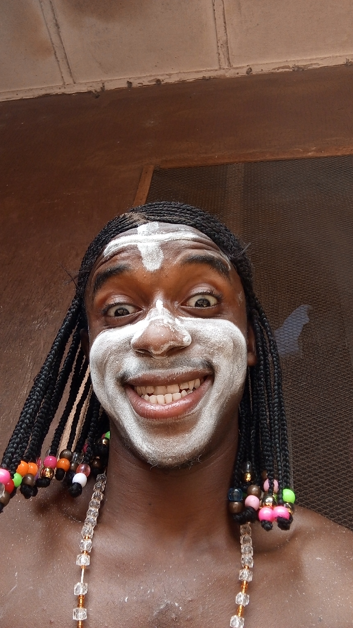 A medium-shot captures a smiling Black man wearing white face paint and a cross on his forehead. The man has braided hair adorned with colorful beads, and he is wearing a beaded necklace. The background shows a doorway covered with a screen. Based on the presence of the Ijele masquerade in other images associated with the location and individual, it is likely that this photograph was taken in Onitsha, Nigeria. The man may be performing a ritual or celebration connected to the masquerade tradition.