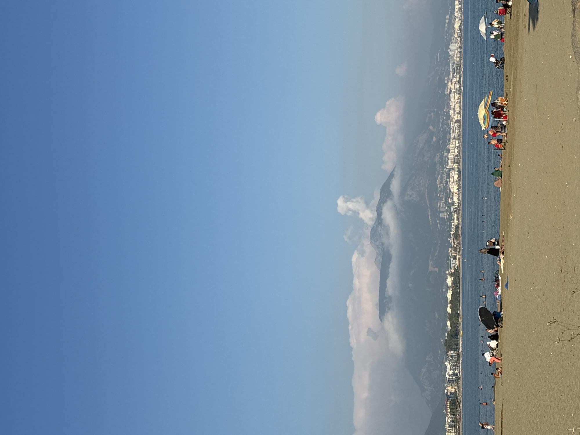 This image captures a sunny day at a beach in Alanya, Turkey, with a clear blue sky and a calm sea. In the foreground, a sandy beach is populated by beachgoers. Some individuals are sitting on the sand under umbrellas, while others are in the water, swimming or wading. There are also groups of people gathered, some appearing to be interacting or relaxing.
Further out, the sea is dotted with people enjoying the water. Beyond the sea, a city skyline is visible, characterized by buildings of varying heights, suggesting a bustling urban area. In the background, majestic mountains rise, partially shrouded by soft, white clouds. The overall atmosphere is one of leisure and enjoyment, typical of a beach destination.