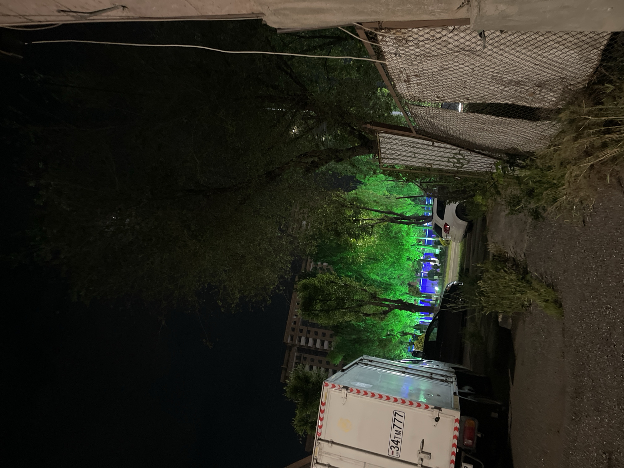 The photograph shows an alleyway in Yerevan, Armenia at night. A white delivery truck with the license plate "34TM777" is parked along the alleyway. Beyond the truck, illuminated green trees line the street. A high-rise building is visible in the background. A chain-link fence runs along one side of the alley.