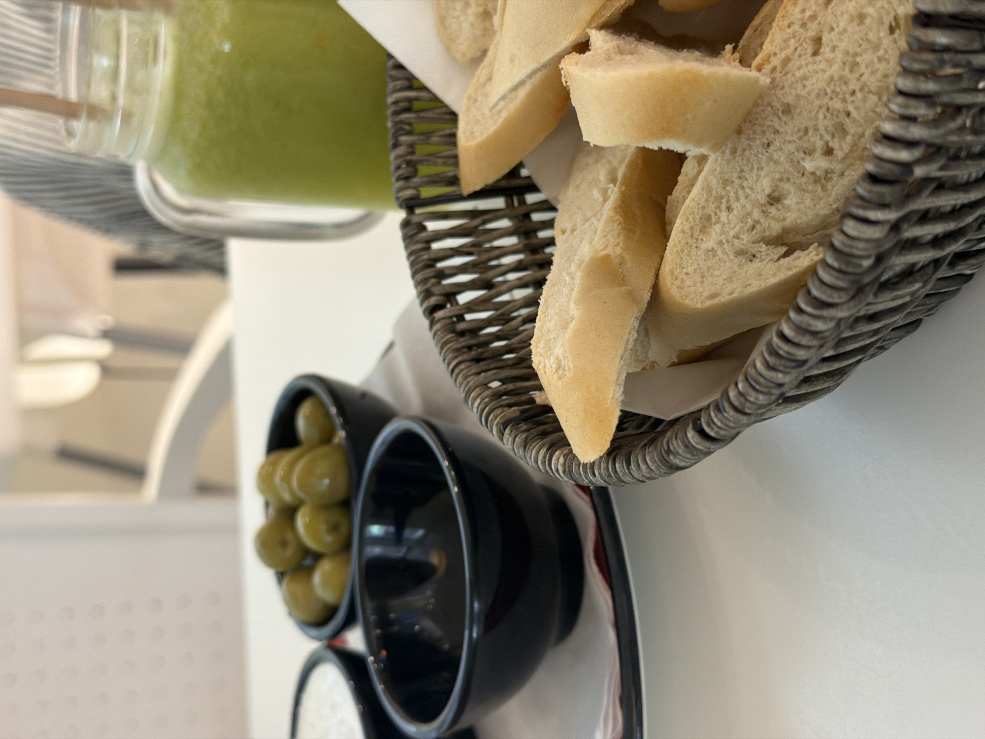 The image shows a close-up of a table setting, likely in a restaurant or cafe. The main objects in focus are a glass jar filled with a green smoothie or juice, a woven basket filled with sliced baguette bread, and two small black bowls containing green olives and possibly a dip or sauce.
The smoothie appears vibrant green and is in a clear glass jar with a wooden stirrer. The baguette slices are golden brown and appear fresh, resting in a dark woven basket lined with white paper. One black bowl is filled with plump green olives, while the other is empty, revealing a glossy black interior.
The background is blurred, but suggests an indoor setting with white tables and chairs. The lighting appears natural, hinting at daytime. The overall presentation suggests a light meal or appetizer service.
There is no visible text in the image. The scene is well-lit and the focus is sharp on the food items, indicating a deliberate composition for presentation. The context of Sant Antoni De Portmany, Spain, suggests a Mediterranean setting, which aligns with the olives and bread.