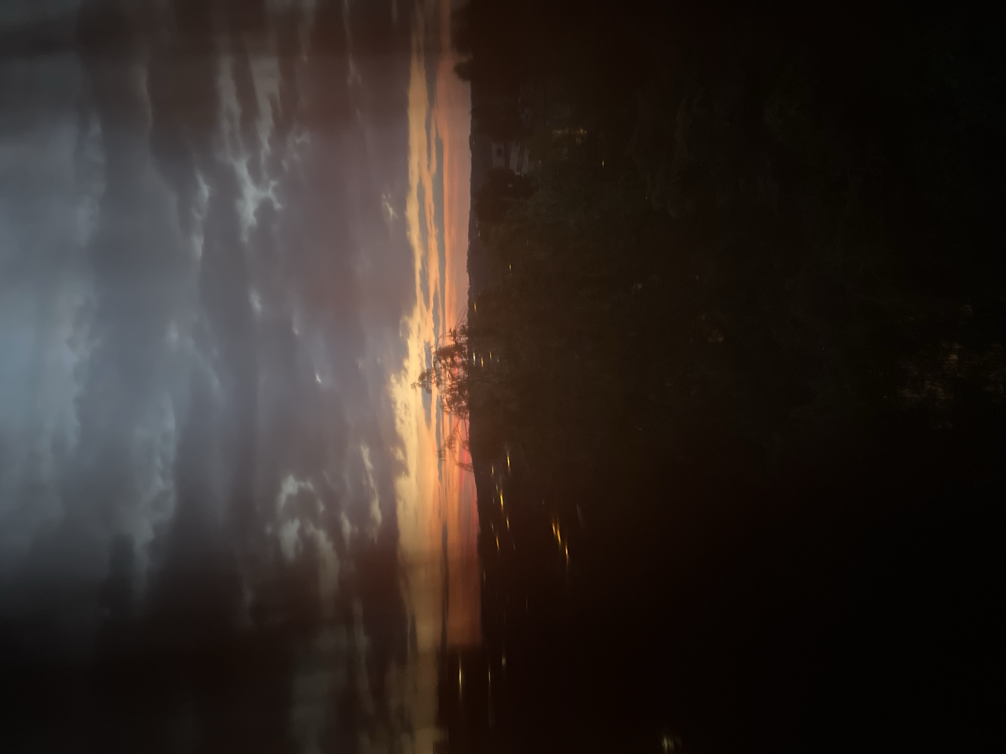 The image depicts a landscape at dusk. The sky is filled with a mix of dark, moody clouds and areas of vibrant orange and pink hues, characteristic of a sunset. The horizon is dotted with lights, suggesting a city or town in the distance. In the foreground, silhouetted trees and foliage obscure much of the ground, adding to the atmospheric and somewhat mysterious quality of the scene. The overall impression is one of a serene, yet dramatic, twilight setting over an urban or suburban area.