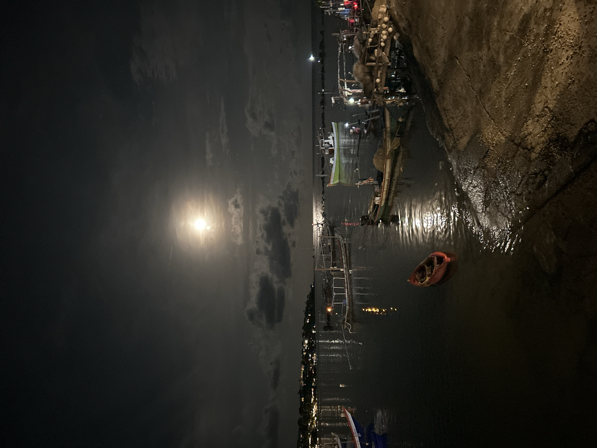 At night in Baan Chaweng Noi, Thailand, a waterfront scene shows boats docked along a concrete quay. Overhead, a bright moon illuminates the water, reflecting light onto the boats and the damp concrete. An orange kayak sits in the dark water. Buildings with lights dot the shoreline in the distance.
