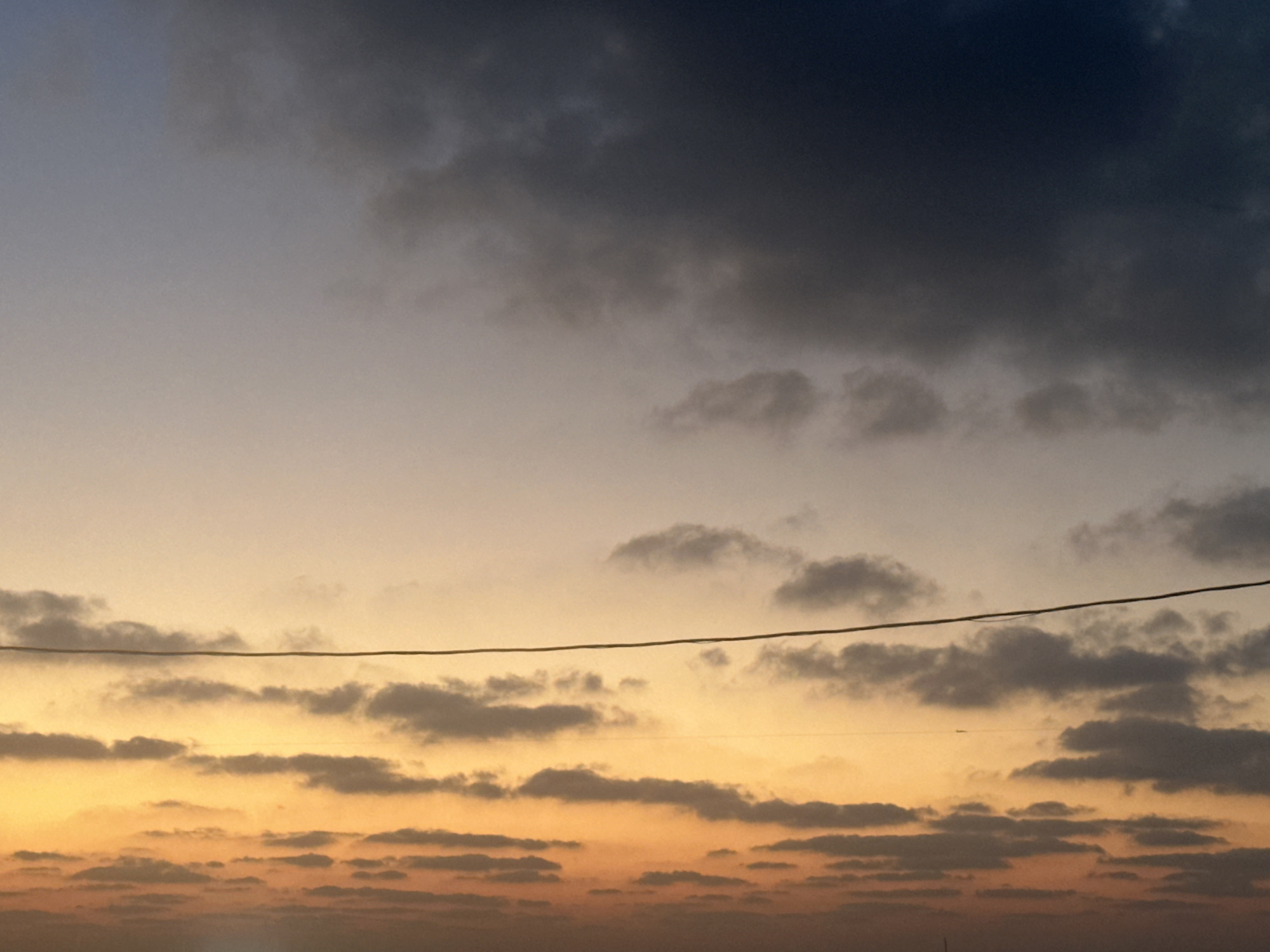 The image captures a dramatic sunset over the city of Haifa, Israel. The sky is a canvas of changing colors, transitioning from a deep, dark blue at the top to a warm, vibrant orange and yellow near the horizon. Scattered clouds, ranging from dark and ominous to wispy and light, populate the sky, adding texture and depth to the scene.

Stretched horizontally across the mid-section of the image is a thin, dark wire, likely an electrical or telephone line, that bisects the sky and the clouds. This wire serves as a stark, man-made element against the natural beauty of the sunset. In the distance, the faint silhouette of the cityscape of Haifa can be observed, subtly merging with the lower part of the image where the colors are most intense. There are no people or discernible actions depicted in the scene. The overall impression is one of a tranquil yet awe-inspiring natural phenomenon occurring within an urban setting.