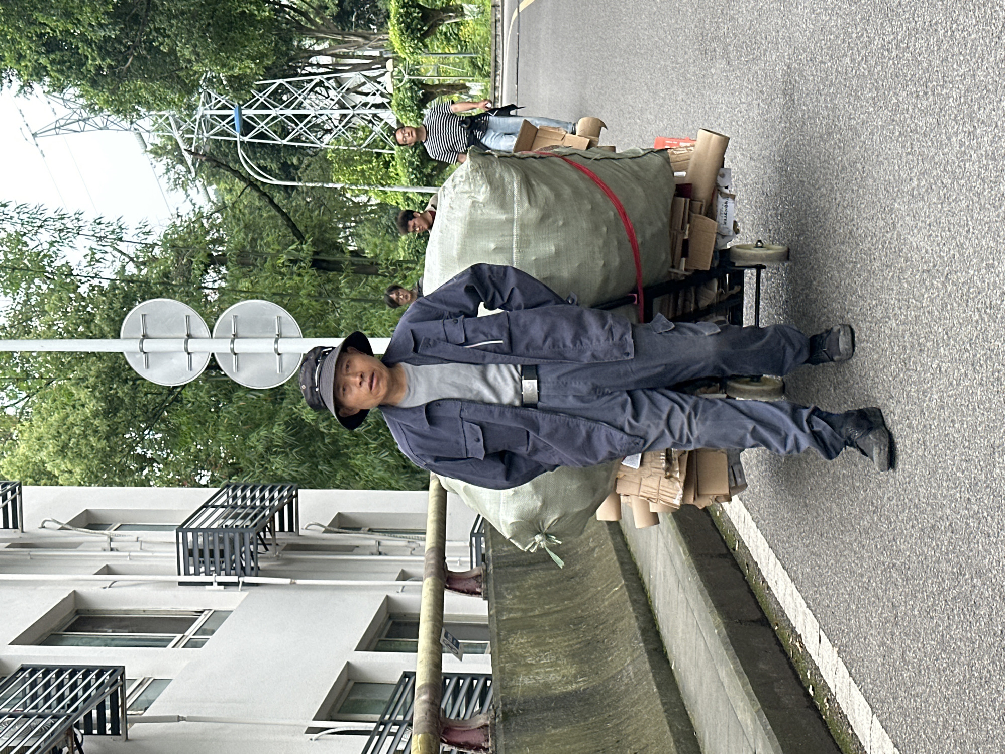 A man wearing a blue collared shirt, blue pants, black shoes, and a hat pulls a two-wheeled cart containing a large green bag and cardboard boxes along the side of a paved road. Several other people are standing in the background. A building with window air conditioning grates is on the left side of the frame. A street sign is to the left of the man. Trees are visible in the background. In the far background, power lines are seen. This scene is in the city of Shapingba District, China.