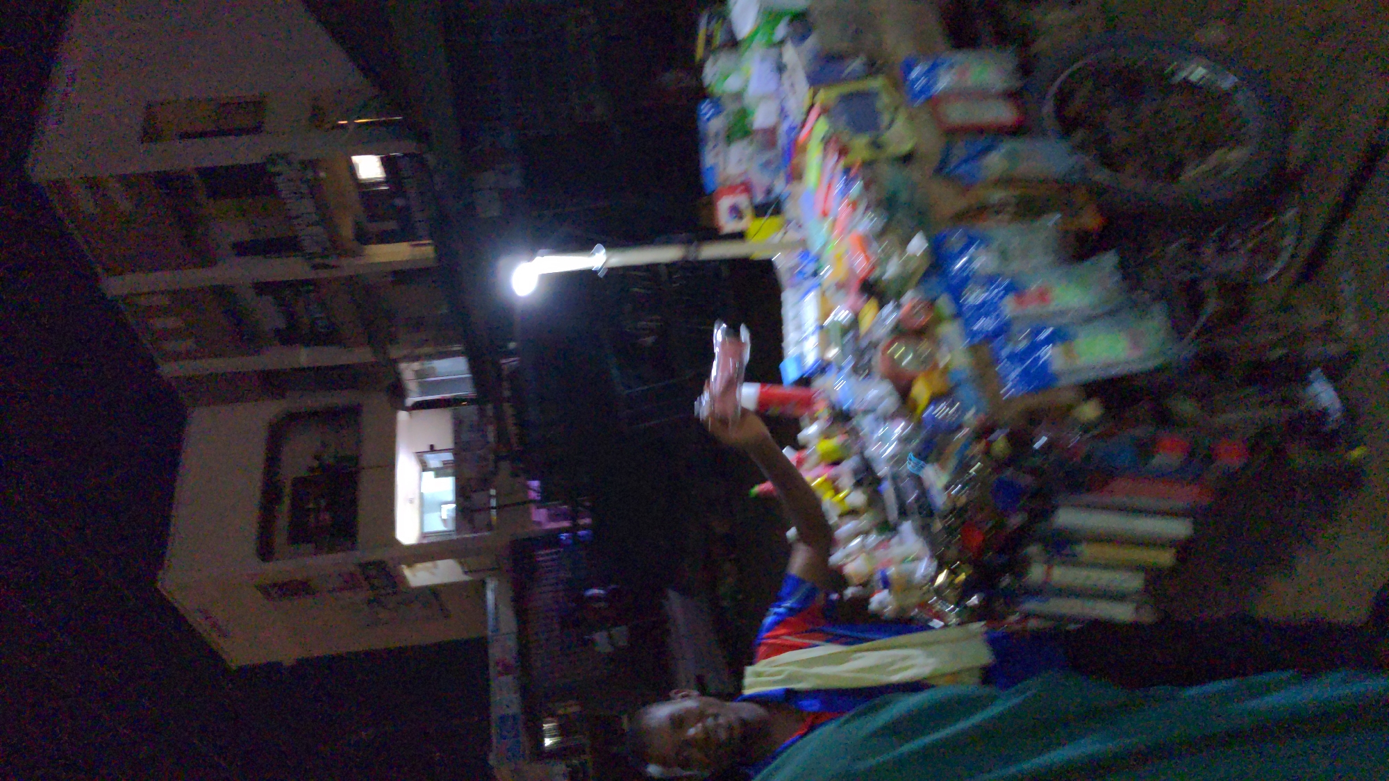 A man lies on a green cloth in Ado, Nigeria, holding a pink plastic bottle in his right hand. He is next to a table laden with various items, including blue packages, clear bottles, and small multicolored objects. A bicycle wheel leans against the table's edge. Behind him, a street light illuminates a multi-story building with visible windows.