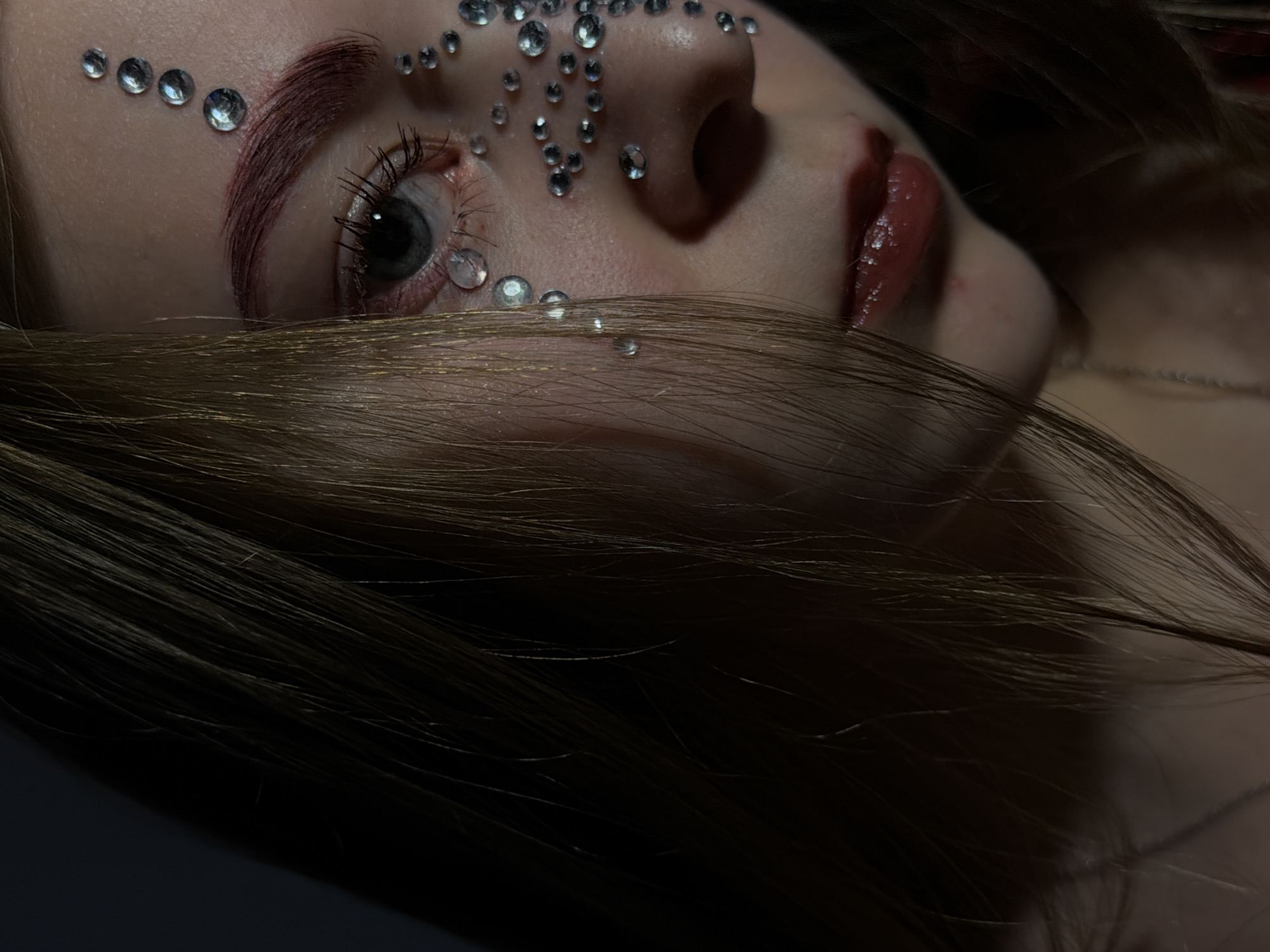 Close-up, angled view of a person's face adorned with decorative rhinestones. The person is lying down, their head tilted back slightly. Rhinestones are placed on the forehead, around the eye, and along the nose bridge. The person has a single visible eye with long lashes and a dark pupil. Red lipstick is visible on the lips. A strand of light brown hair partially covers the lower part of the face. The photograph has low lighting. There are no discernible landmarks to confirm the location in Perm, Russia.