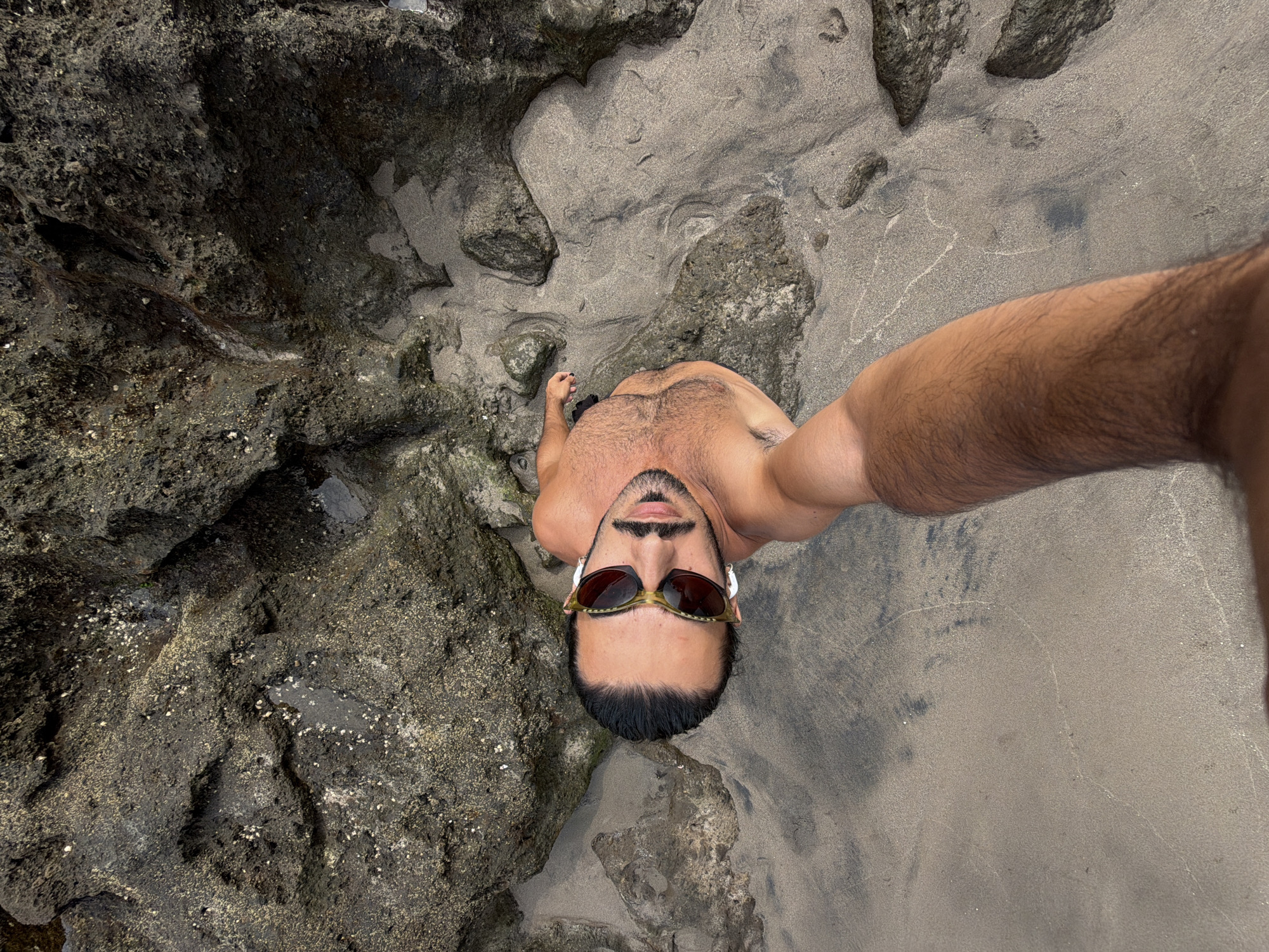 A high-angle, wide shot captures a shirtless man with dark hair and sunglasses resting on a sandy beach in North Kuta, Indonesia. The man is positioned in the center of the frame, looking upwards towards the camera, his right arm extended to hold the camera and take the selfie. He has a mustache and goatee, and is wearing white earbuds. The beach is characterized by dark, porous volcanic rocks and wet sand, with some areas appearing to be tide pools. The lighting suggests it is daytime.