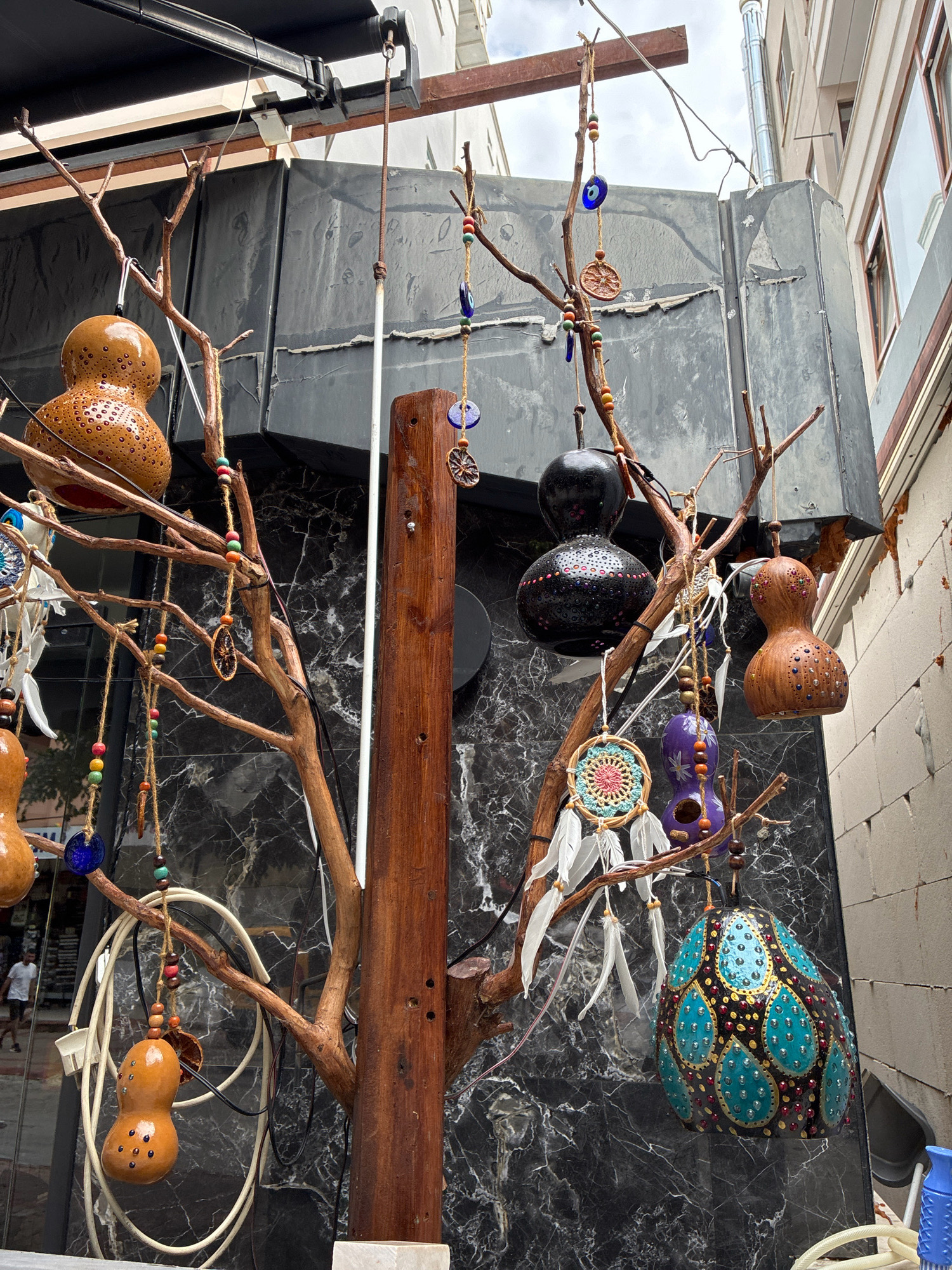 The image shows a street scene in Alanya, Turkey, focusing on a decorative tree made of branches with various ornaments. The tree is adorned with dried gourds, some painted with intricate patterns, and others decorated with beads and charms. Among the ornaments are blue "evil eye" talismans, dreamcatchers, and dried fruit slices. The backdrop consists of a building with a dark, marbled facade and a white brick wall. Several electrical cords are also visible, coiled on the ground and near the tree. In the background, a person is walking on the street. The lighting suggests it is daytime.