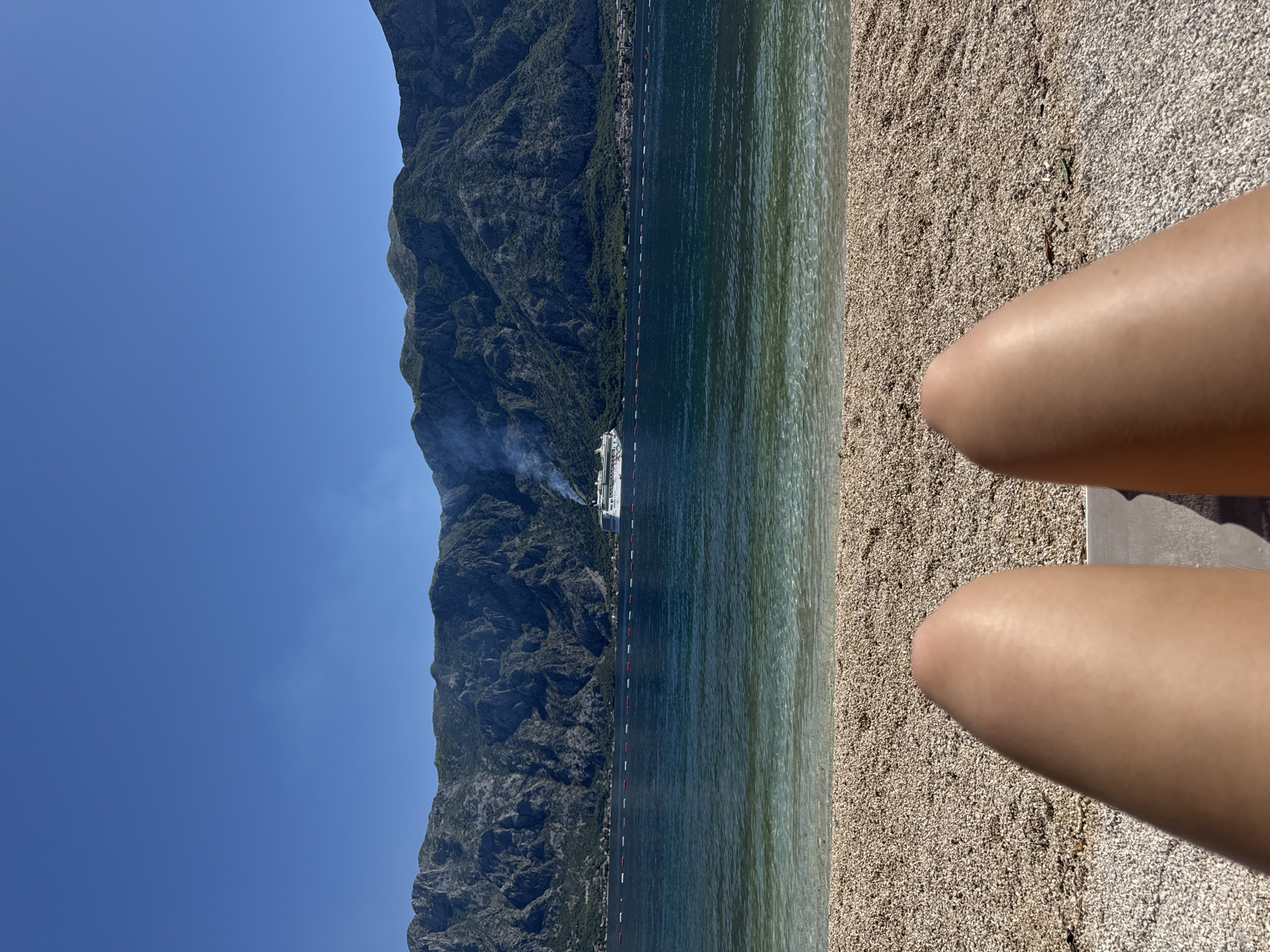 The image shows a view from a beach in Prčanj, Montenegro. The foreground contains a small section of the beach and the legs of a person who is seated. The beach is composed of small pebbles. Beyond the beach is the calm, clear sea. A cruise ship can be seen in the distance, with smoke billowing from its exhaust stacks. The backdrop is dominated by a large, imposing mountain range with dense greenery. The sky above is clear and blue.