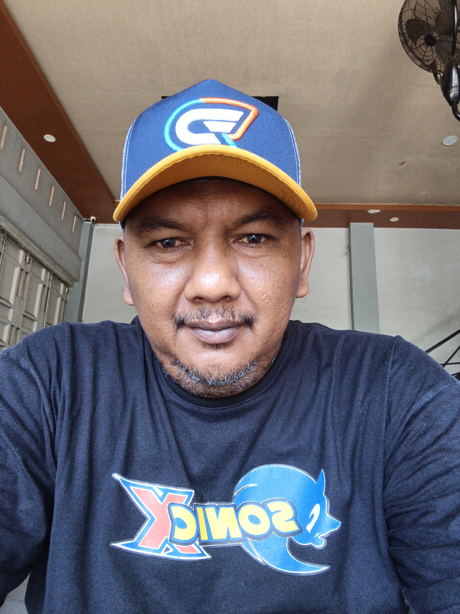 A close-up, outdoor, eye-level selfie shows a man wearing a black t-shirt and a blue and yellow baseball cap. The man has brown skin, a goatee, and is looking directly at the camera with a neutral expression. The t-shirt has a graphic print on it, appearing to be the character Sonic the Hedgehog. The background is out of focus but suggests an outdoor environment with a building and a fan mounted on a ceiling. The overall impression is that of a casual portrait.