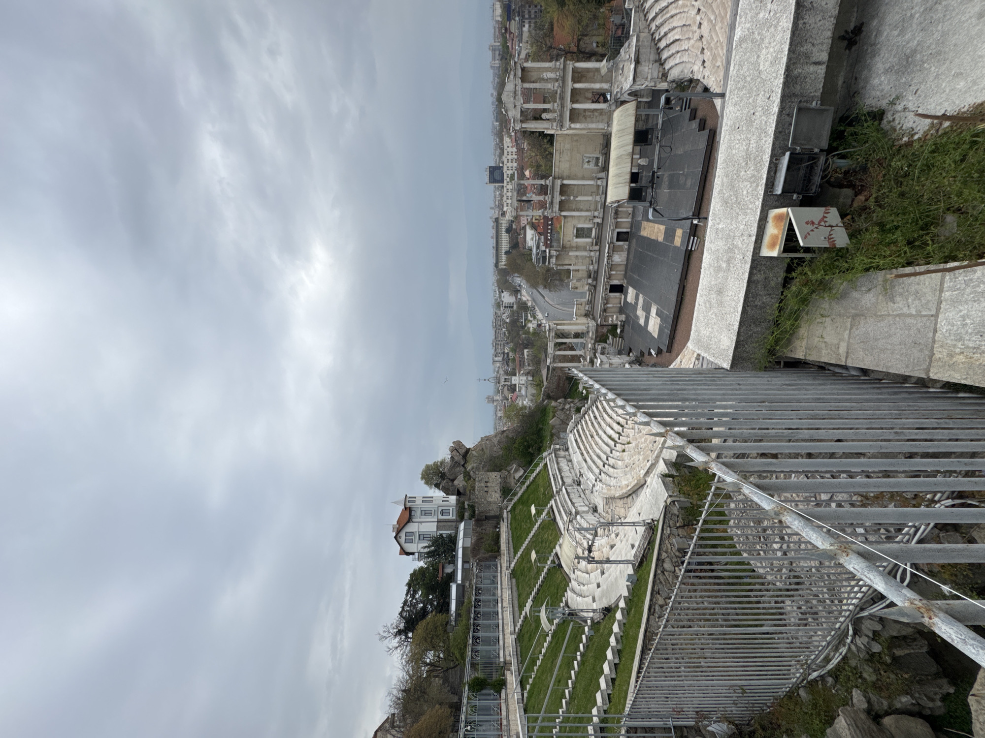 The image shows a high-angle view of the Ancient Theatre of Philippopolis in Plovdiv, Bulgaria, with a section of the surrounding city visible in the background under a cloudy sky. The theater's seating and stage area are prominent, as is a metal railing in the foreground. Nearby structures and vegetation can be seen in the vicinity of the theater.