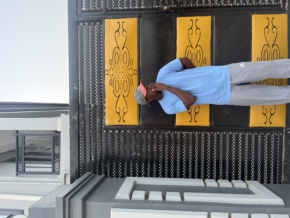 A man stands in front of an ornate gate. He is wearing a light blue shirt, gray pants, and a cap. He has one hand up to his face. The gate is black with yellow rectangular panels decorated with a symmetrical design. The gate is made of vertical black bars punctuated with small white dots. The lower part of the image shows a gray concrete structure with a geometric pattern. A building is visible in the background to the left.