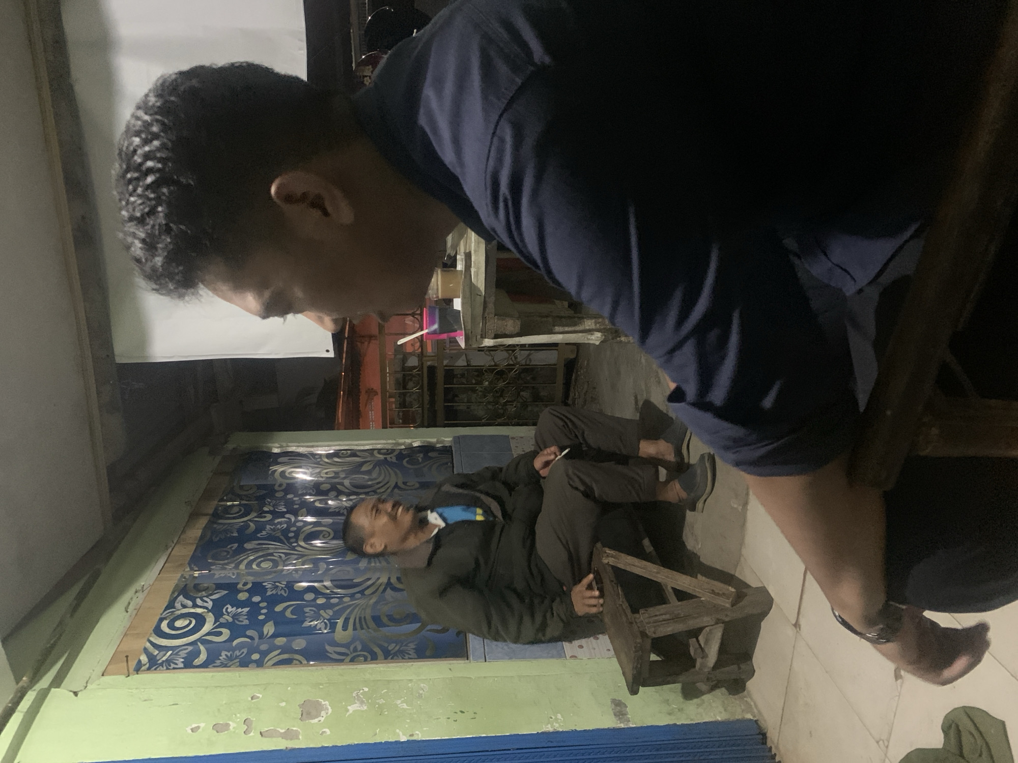 Two men are sitting outside at night. The man on the left is seated on a wooden stool and is wearing a dark jacket over a blue and yellow shirt, and dark pants. He is holding a cigarette in his right hand and smiling. He has short, dark hair. The man on the right is seated on a chair, facing away from the camera and looking to his left. He is wearing a dark blue shirt and has short, dark hair. In front of the man on the right is a small table with two cups, one containing a light-colored liquid. The background shows a building with a green painted wall and blue curtains with a white floral pattern. There is also a white screen hanging on the wall. The ground is tiled. The scene appears to be in a residential area or a small shop in Palembang, Indonesia.