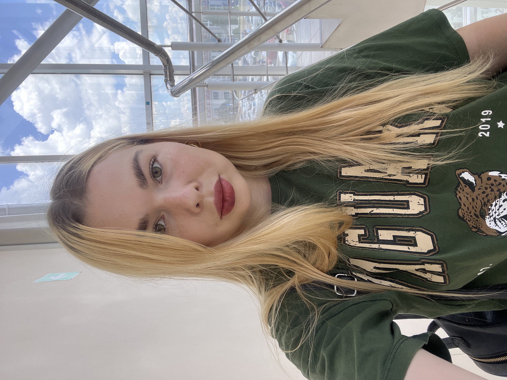 This is a portrait of a young woman with blonde hair and light-colored eyes. She is wearing a dark green t-shirt with a graphic print that includes the text "2019" and what appears to be a leopard's head. Her lips are colored with a dark lipstick. The woman is looking directly at the camera, and her expression is neutral.
The background suggests an indoor location with large windows that offer a view of the sky and some buildings. The sky is bright blue with white clouds, indicating it is daytime and likely good weather. Metal railings and structural elements of the building are visible, suggesting a modern interior, possibly a mall or a public building with a glass facade. A small, light blue and white sign with an arrow is affixed to the wall in the lower left portion of the image.
The image is oriented vertically, and the woman's head is positioned at the top of the frame. Her hair cascades down over her shoulder and onto her chest. The lighting appears to be natural, coming from the windows. No specific activity is taking place beyond the selfie being taken.