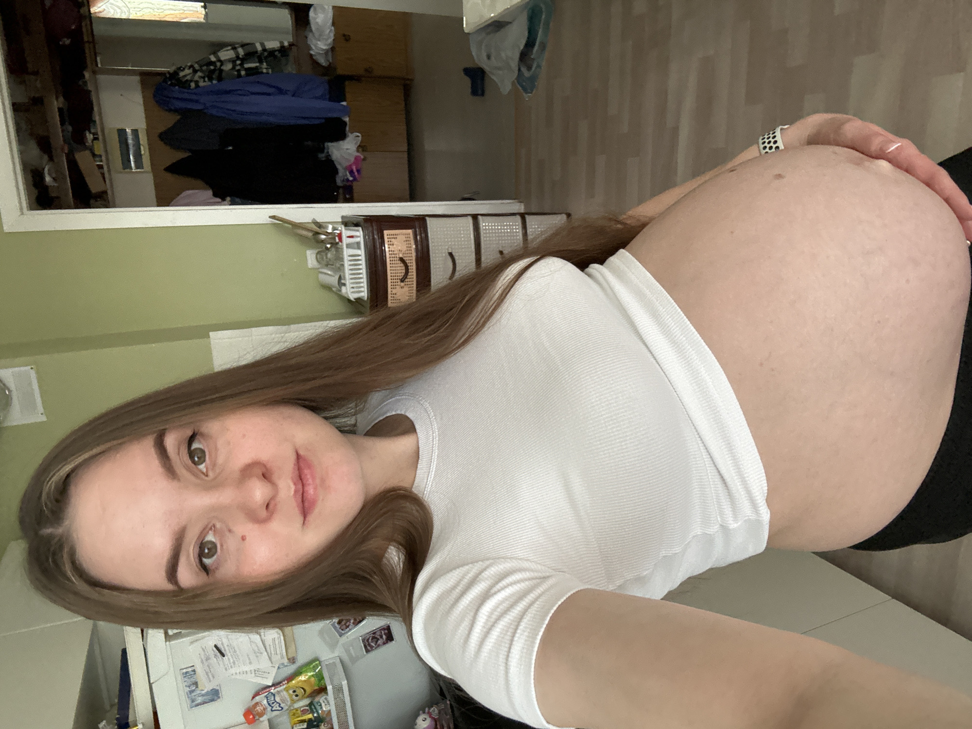 A pregnant person stands in a room, facing the camera and holding their abdomen. They have long brown hair and are wearing a white ribbed t-shirt and black pants. The room appears to be a kitchen or living area with a refrigerator, cabinets, and wooden flooring. In the background, there is an open doorway leading to another room where clothes are hanging and drawers are visible. The overall lighting is natural and soft, indicating it's daytime.